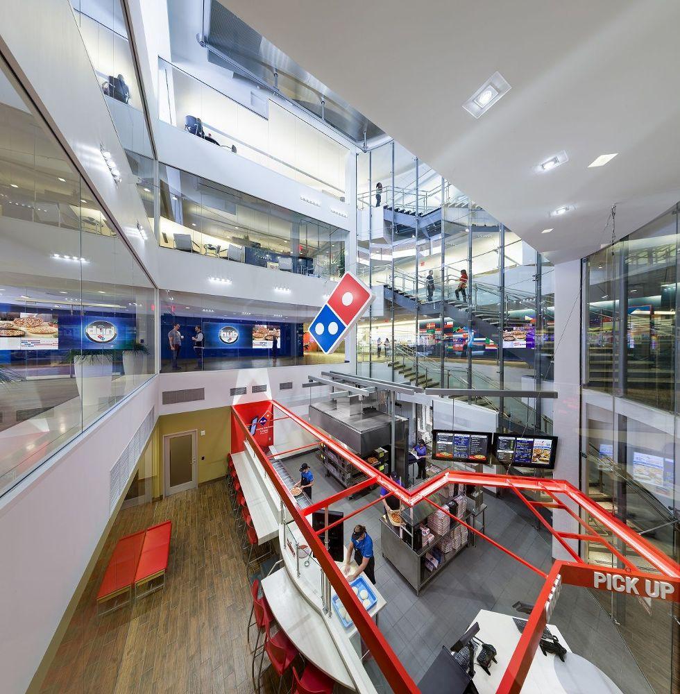 A modern Domino's Pizza store interior featuring multiple levels, large glass windows, and a red pickup area. Staff members are seen working in the kitchen and at the counter.