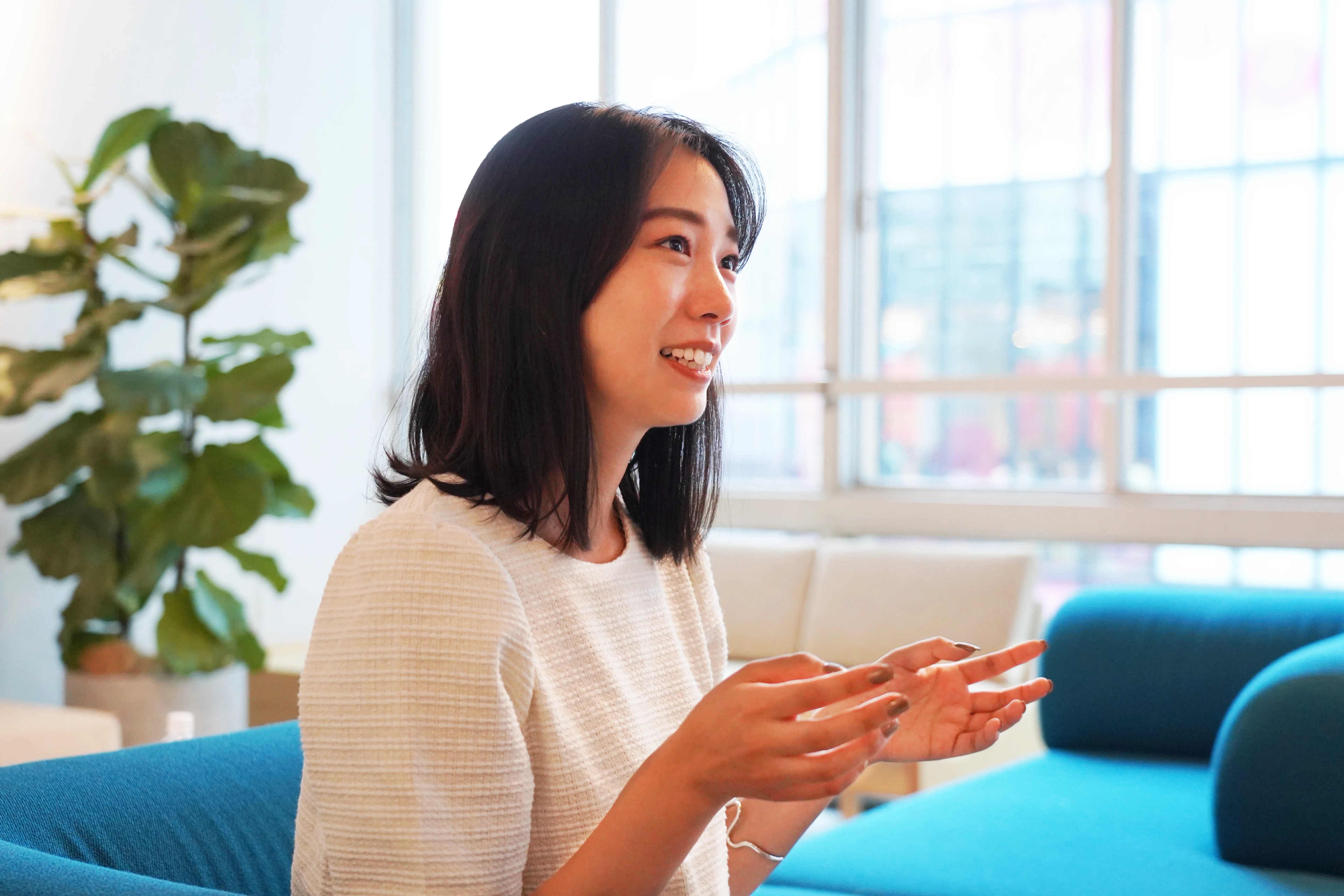 女性がソファに座り、笑顔で話している様子。背景には観葉植物と窓が見える。