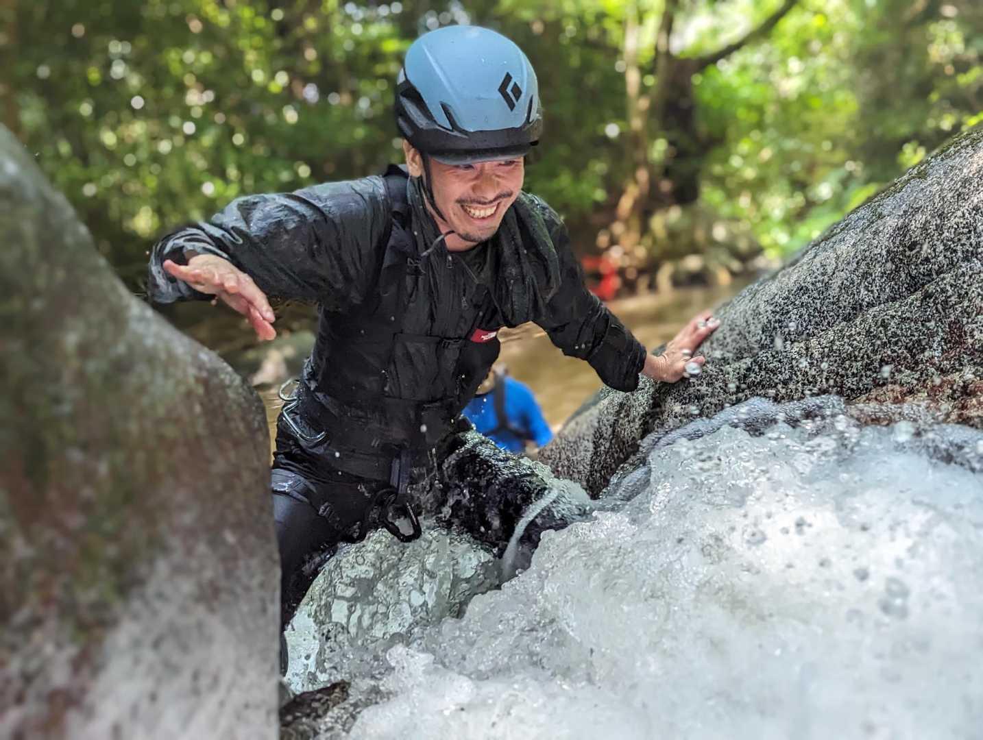 ヘルメットと黒い装備を着けた男性が、笑顔で水しぶきを浴びながら渓流の岩場を登っている。