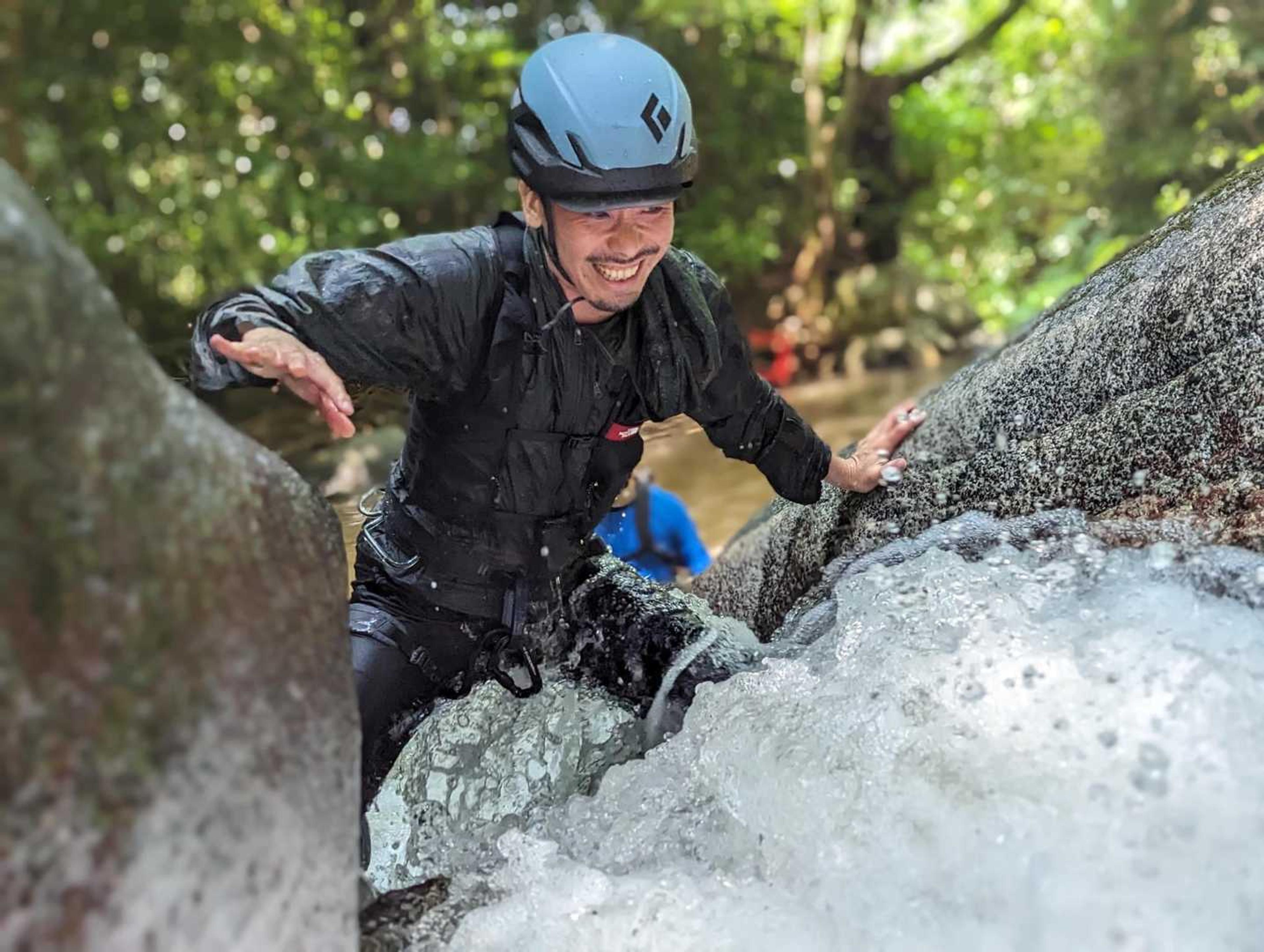 ヘルメットと黒い装備を着けた男性が、笑顔で水しぶきを浴びながら渓流の岩場を登っている。