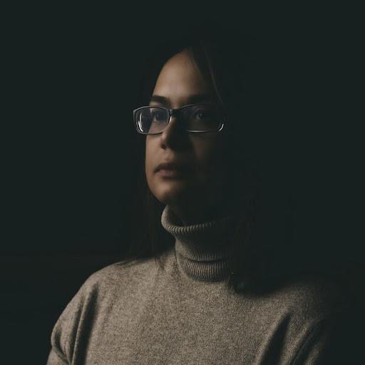 A woman with glasses and a turtleneck sweater is illuminated against a dark background, looking thoughtfully to the side. The lighting highlights her facial features and expression.