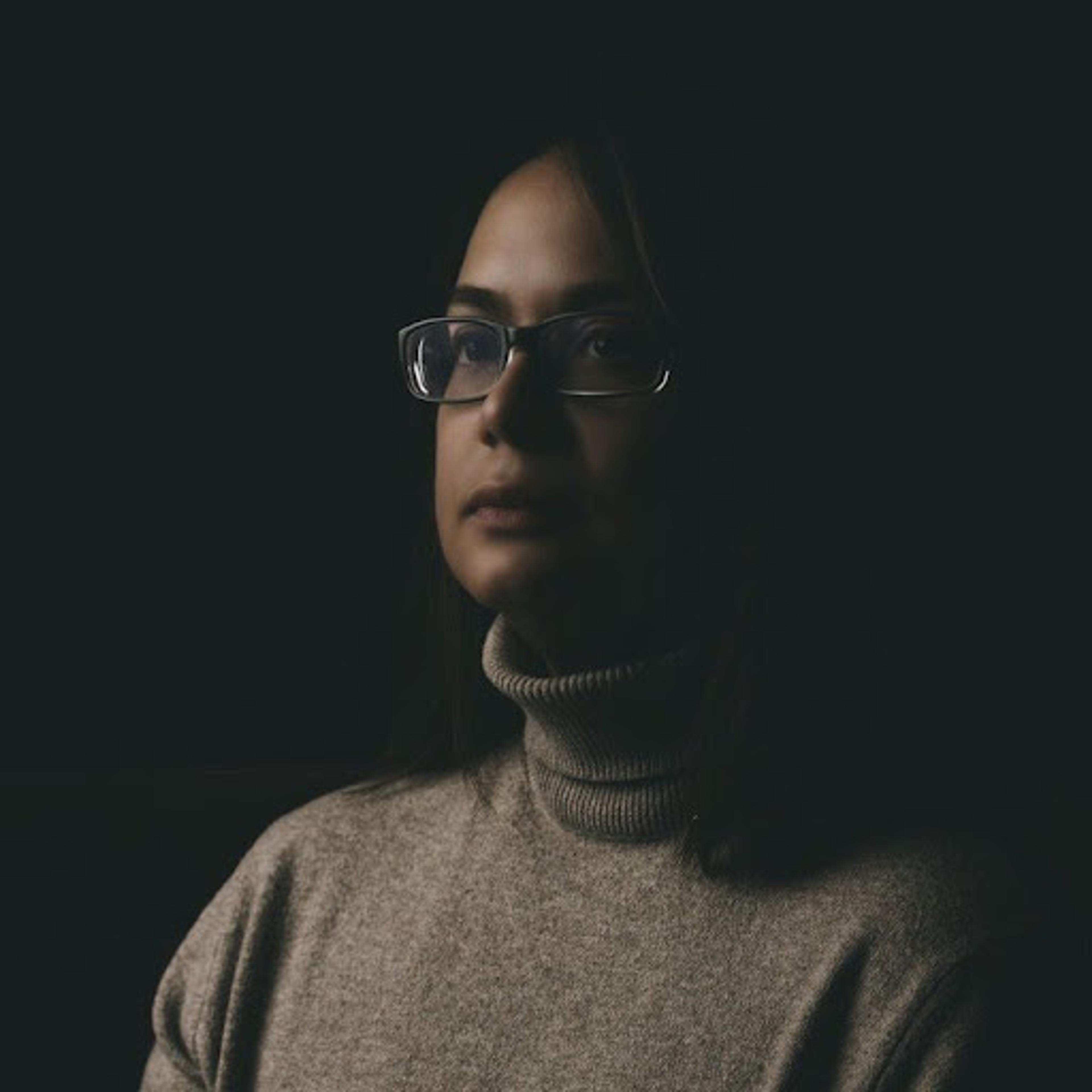 A woman with glasses and a turtleneck sweater is illuminated against a dark background, looking thoughtfully to the side. The lighting highlights her facial features and expression.