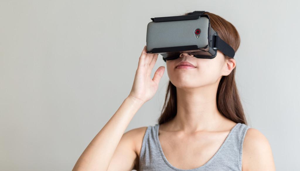 A woman wearing a virtual reality headset smiles while adjusting it. She has long brown hair and is dressed in a gray tank top.