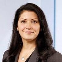 A woman with long dark hair smiles confidently, wearing a gray blazer. She is in a professional setting with a light background.