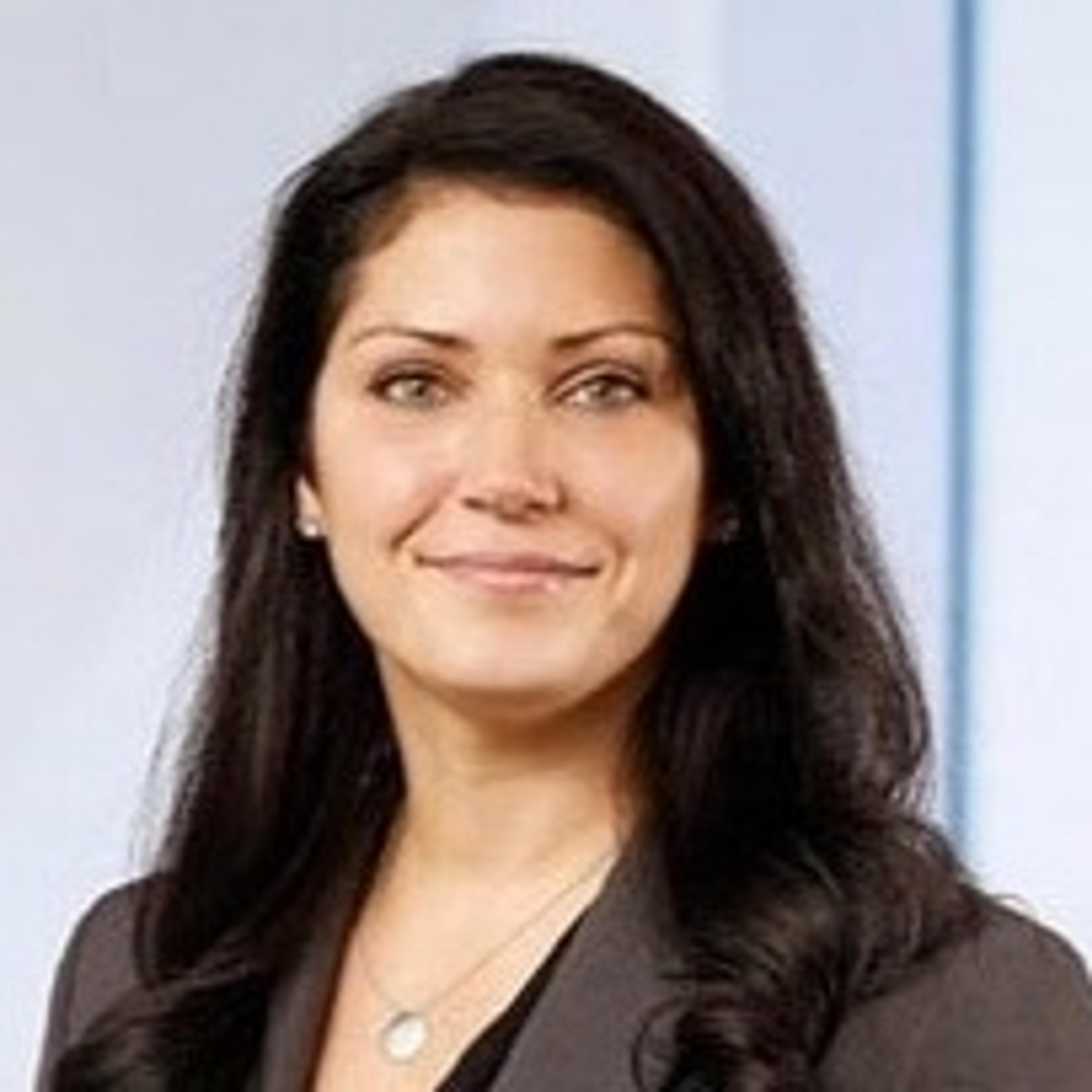 A woman with long dark hair smiles confidently, wearing a gray blazer. She is in a professional setting with a light background.