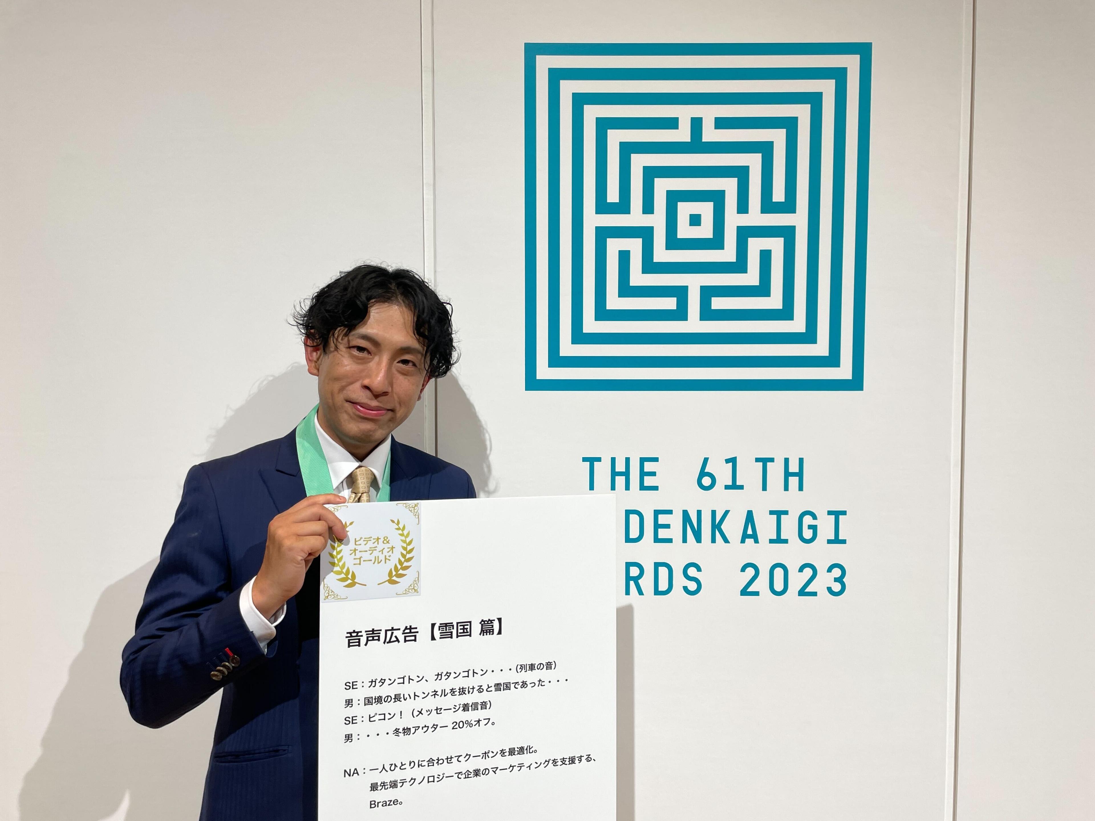 受賞者が「第61回電会議アワード2023」の賞状を持っている写真。背景にはロゴが描かれています。