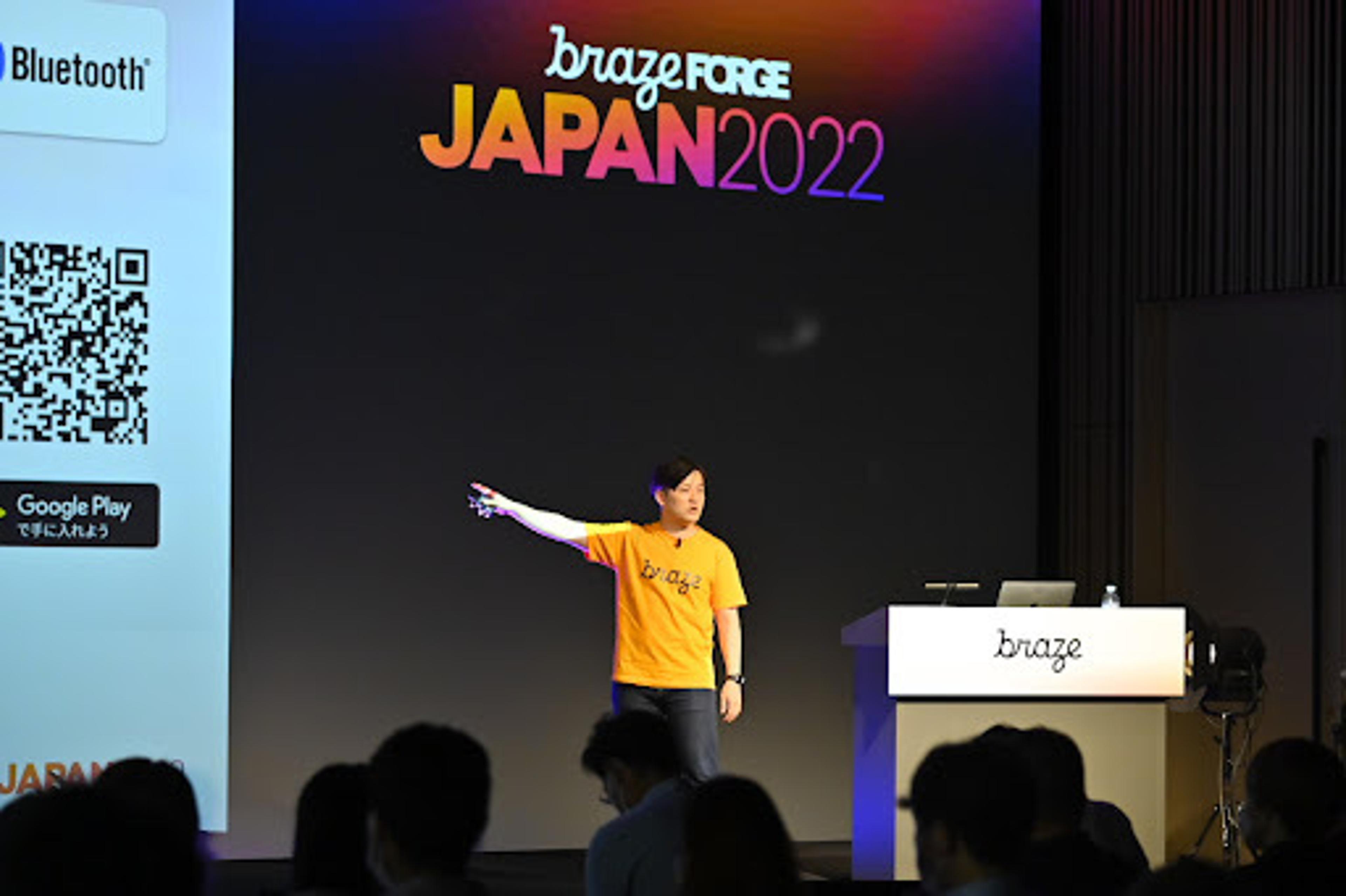 ブラゼフォースジャパン2022のイベントで、オレンジのTシャツを着た講演者がステージで話しています。背景にはカラフルなロゴが表示されています。