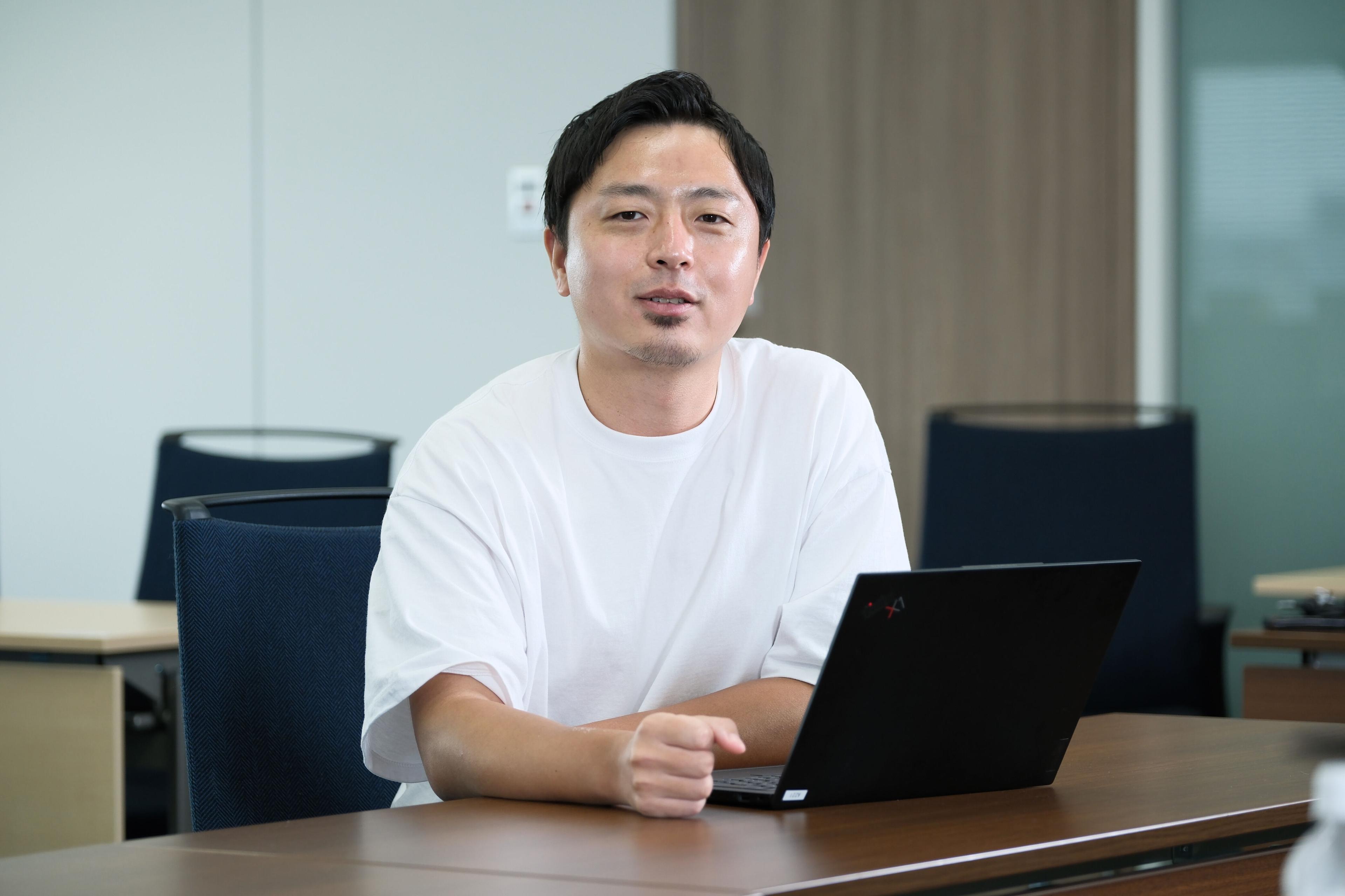 白いTシャツを着た男性がノートパソコンの前に座っています。彼はカメラに向かって話しかけています。
