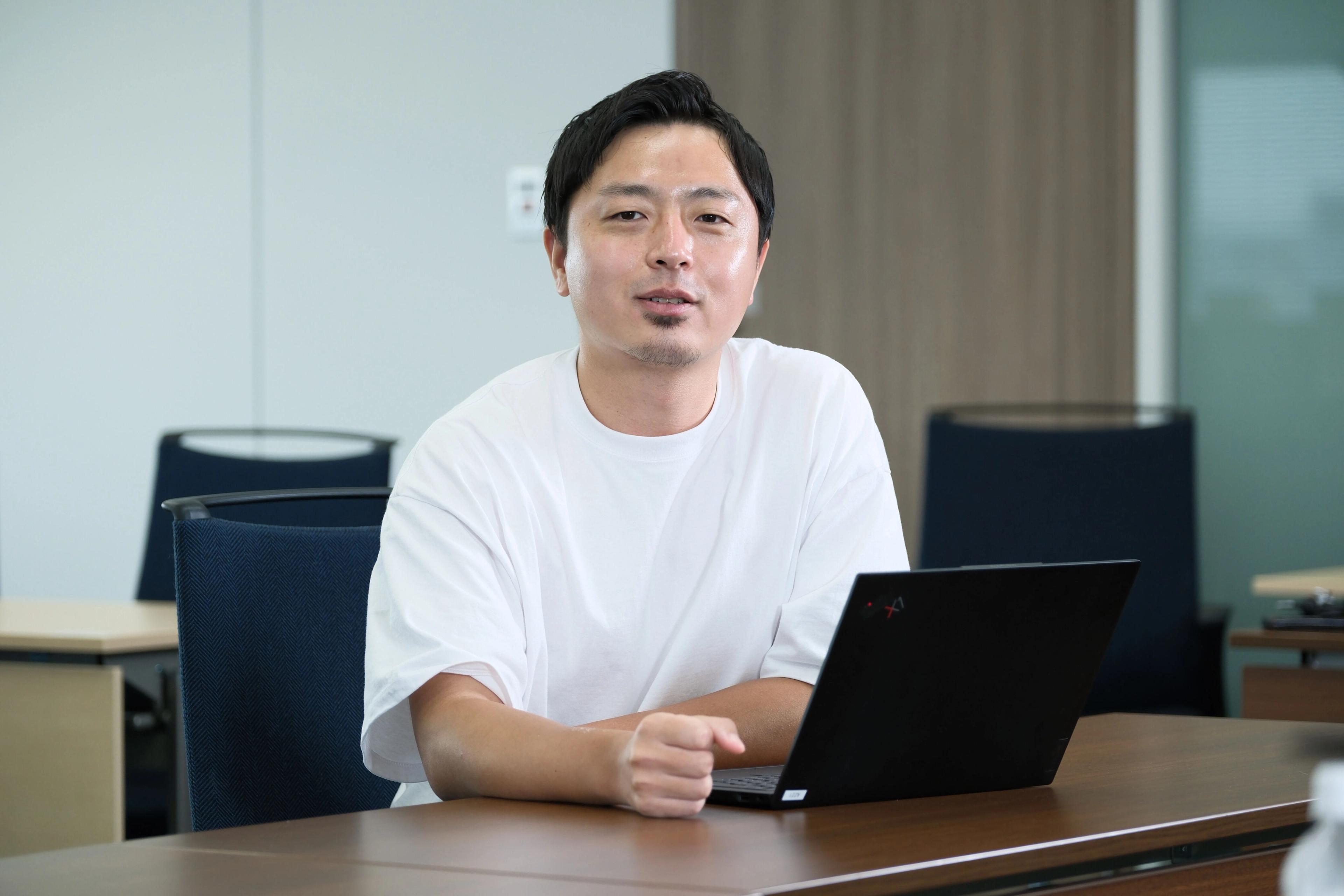 白いTシャツを着た男性がノートパソコンの前に座っています。彼はカメラに向かって話しかけています。