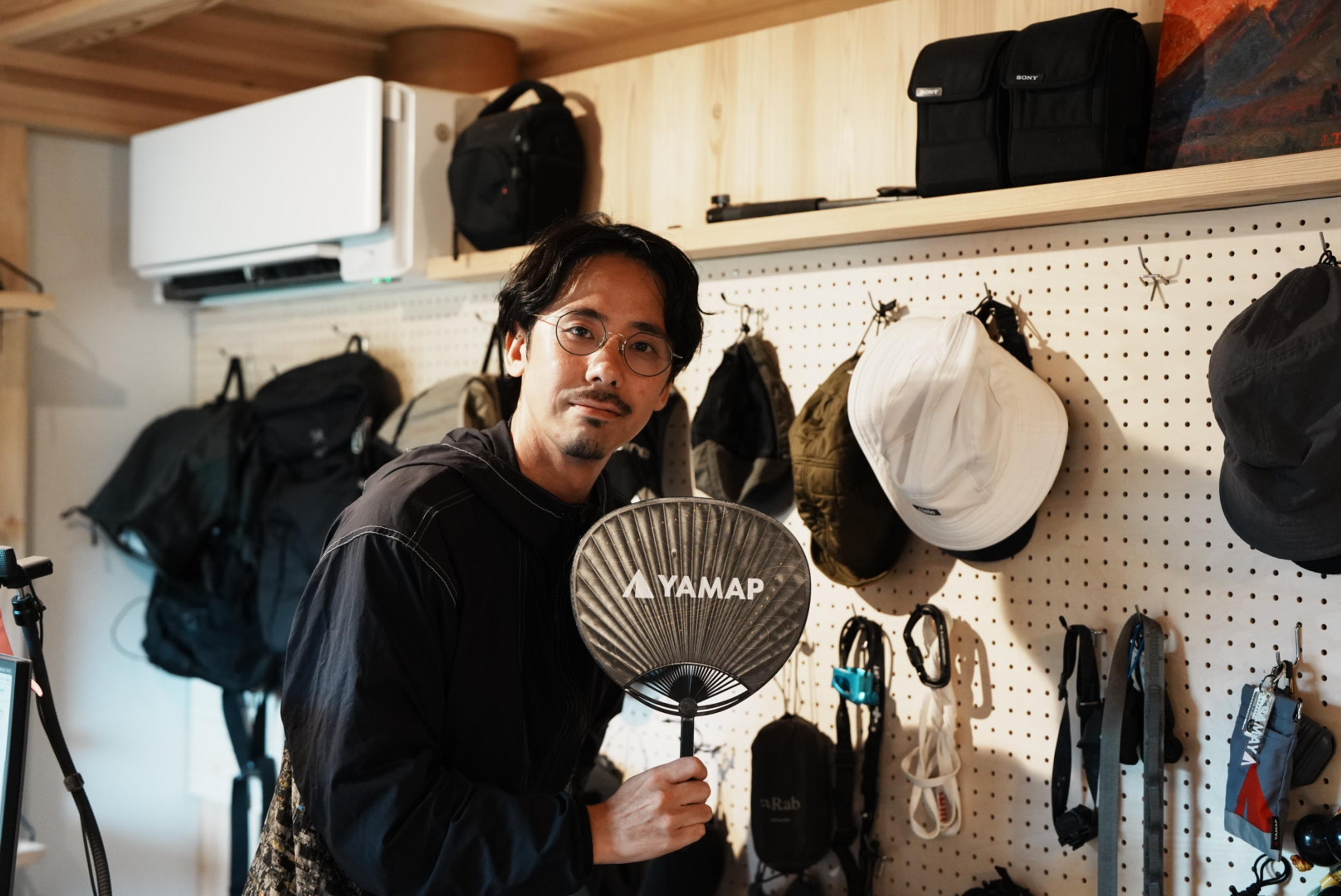 登山用品が並ぶ壁の前で、眼鏡をかけた男性が「YAMAP」と書かれたうちわを手にしている。