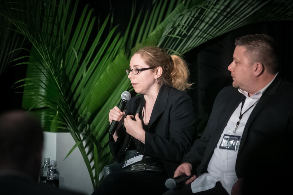 A woman with glasses speaks into a microphone while seated next to a man at a panel discussion, with green plants in the background.