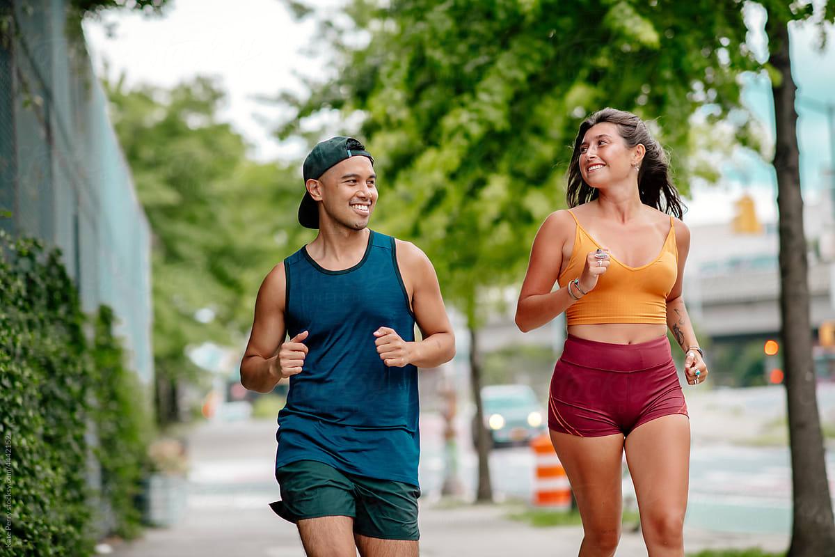 A man and woman jog together on a tree-lined path, both smiling and wearing athletic clothing. The background shows a blurred cityscape.