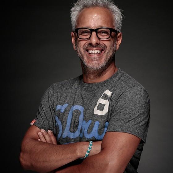A smiling man with gray hair and glasses stands with his arms crossed, wearing a gray t-shirt. The background is dark, highlighting his cheerful expression.