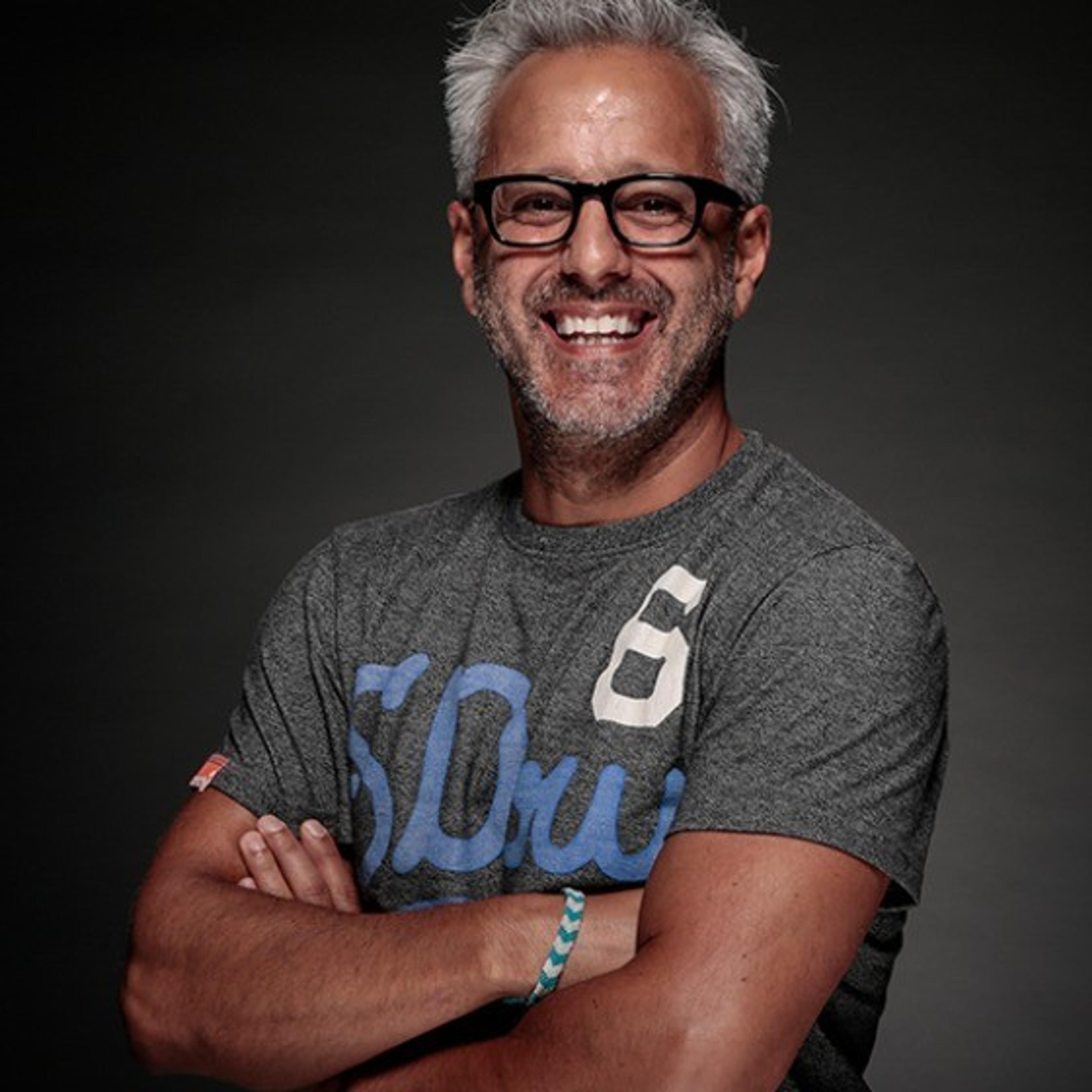 A smiling man with gray hair and glasses stands with his arms crossed, wearing a gray t-shirt. The background is dark, highlighting his cheerful expression.