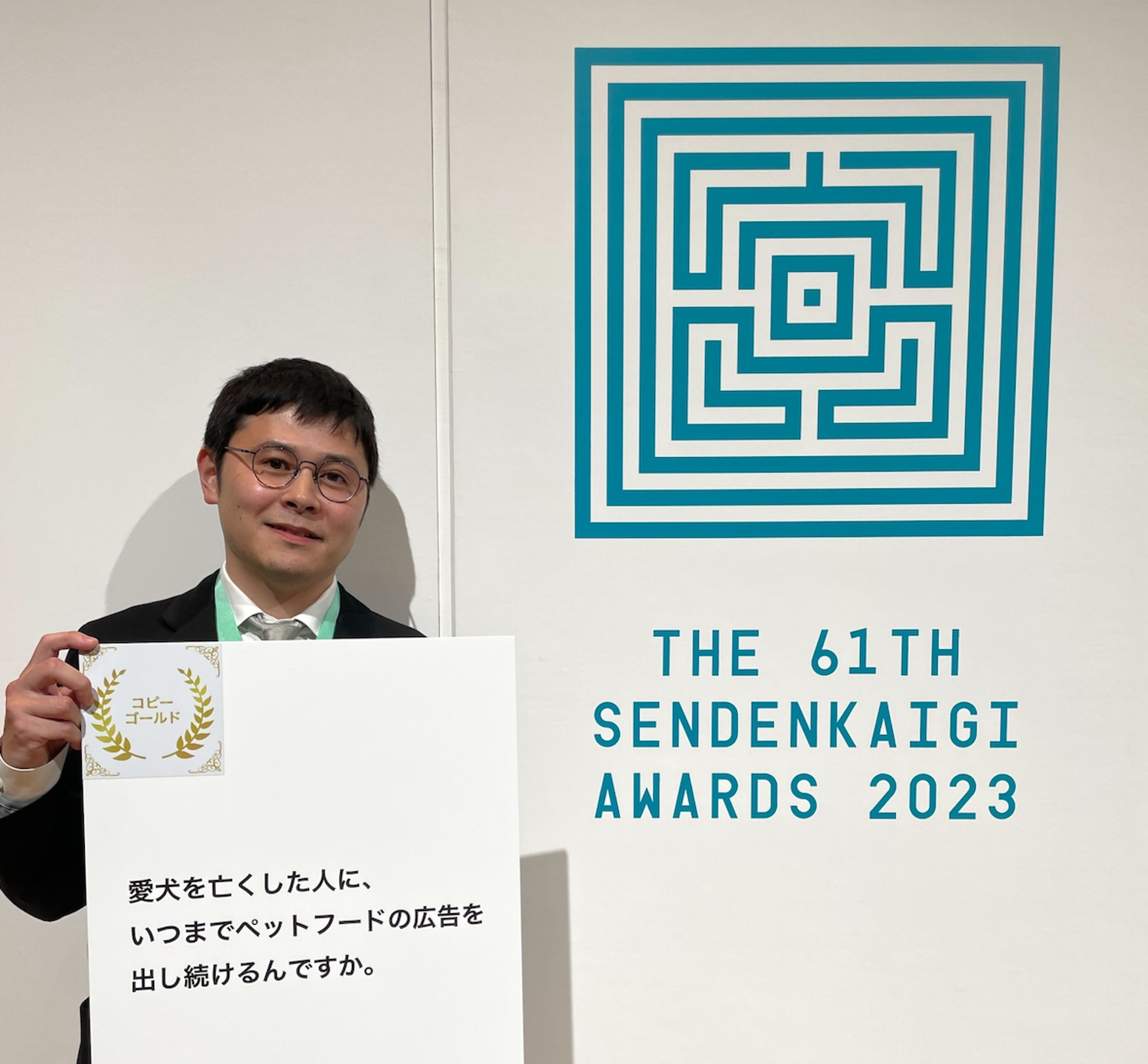 ペットフードの広告を持つ男性が、2023年の第61回宣伝会議アワードの前でポーズをとっています。背景にはロゴがあります。