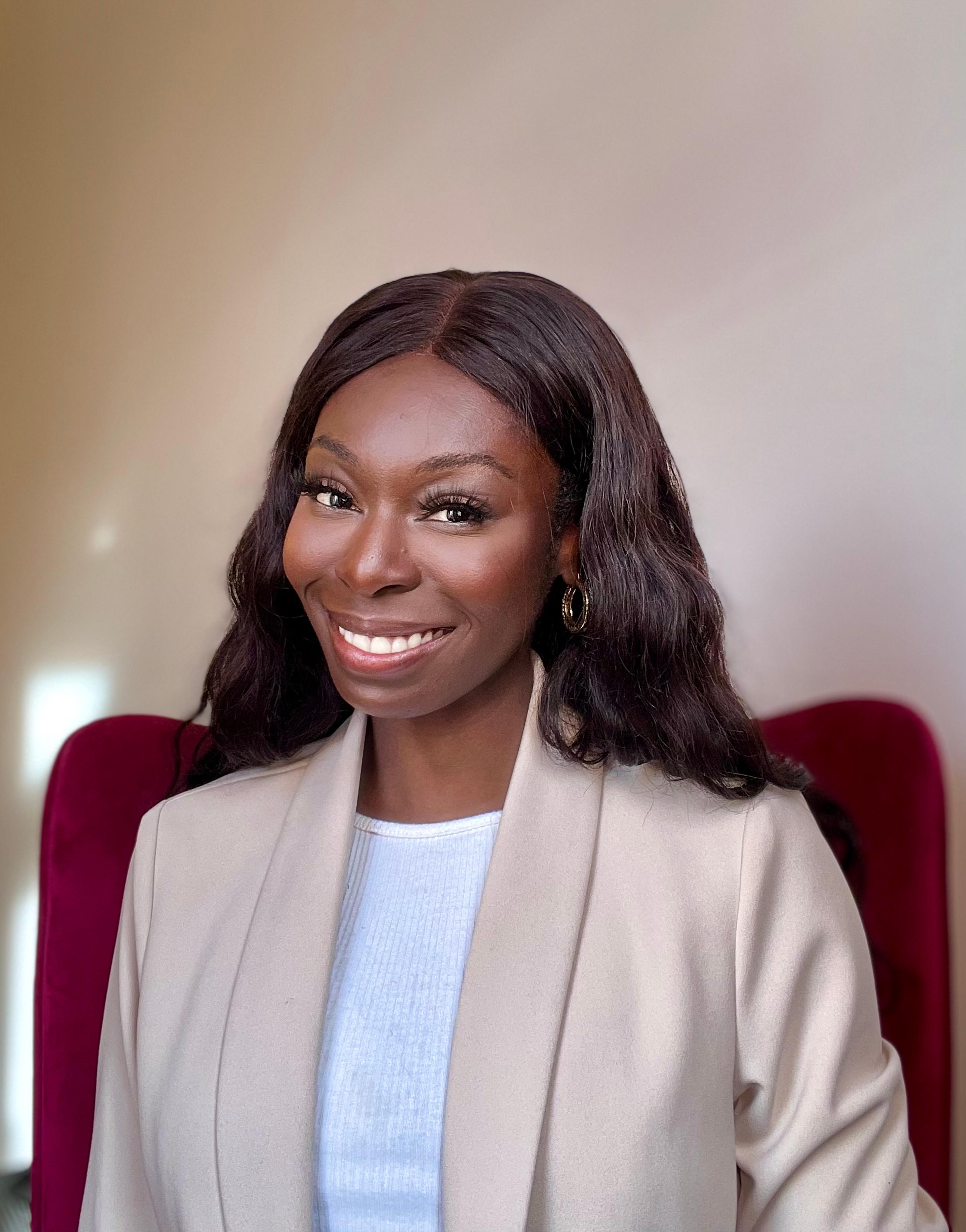 A woman with long, wavy dark hair smiles while wearing a light-colored blazer over a white top. She is seated against a soft, neutral background.