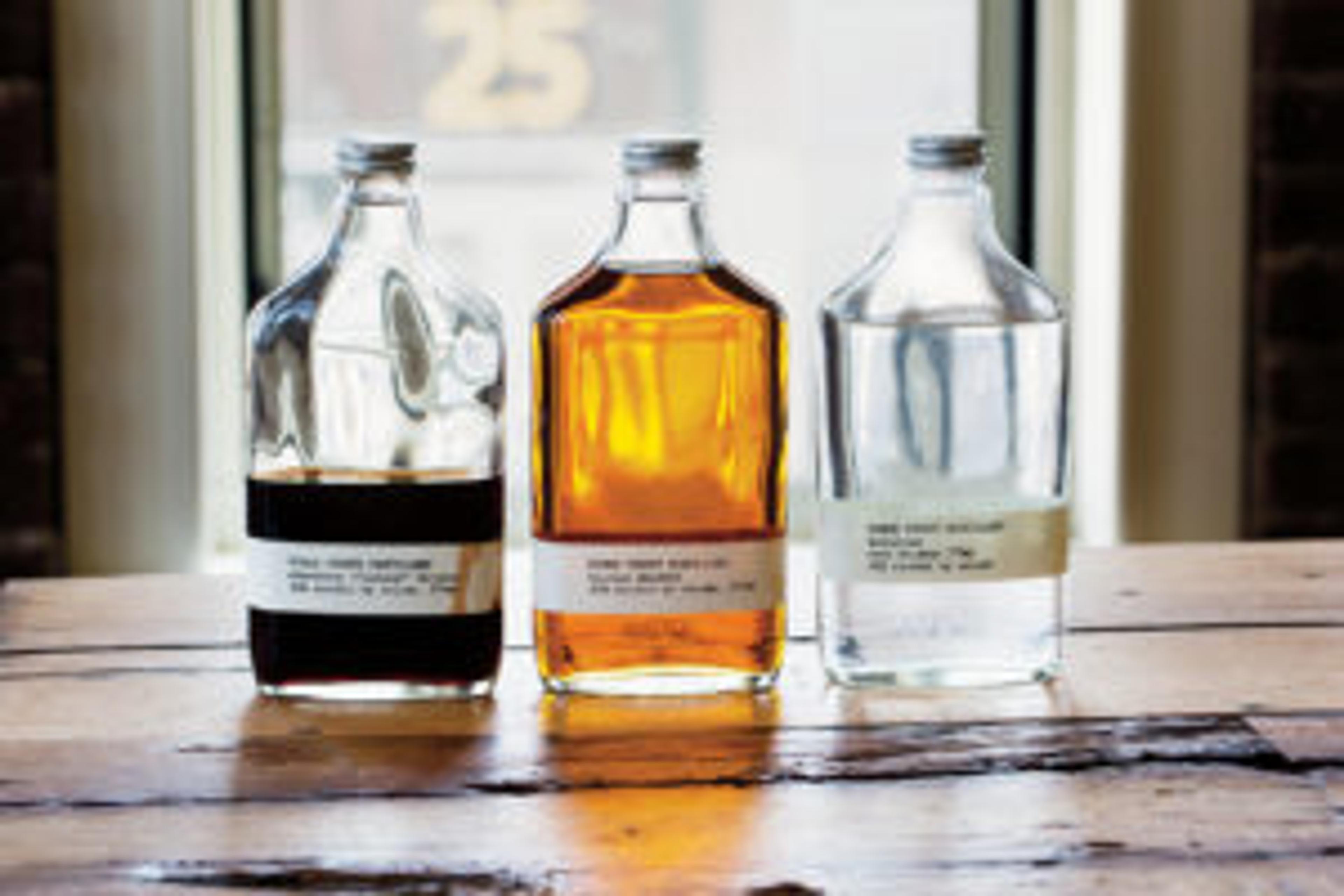 Three glass bottles filled with different liquids sit on a wooden table. The left bottle contains a dark liquid, the middle bottle has a golden liquid, and the right bottle holds a clear liquid.