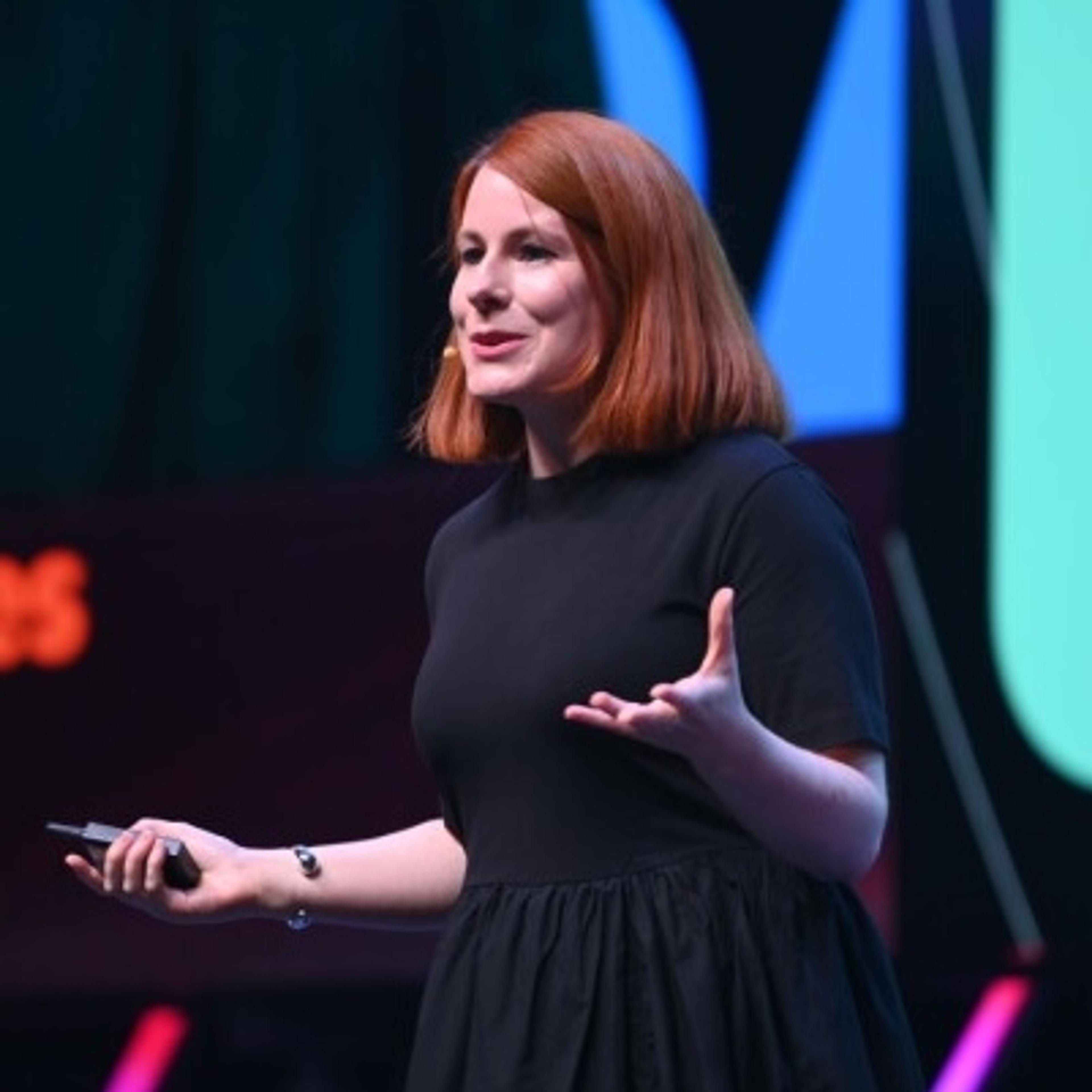 A woman with shoulder-length red hair speaks on stage, gesturing with one hand while holding a microphone in the other. She is wearing a black dress and appears engaged with the audience.