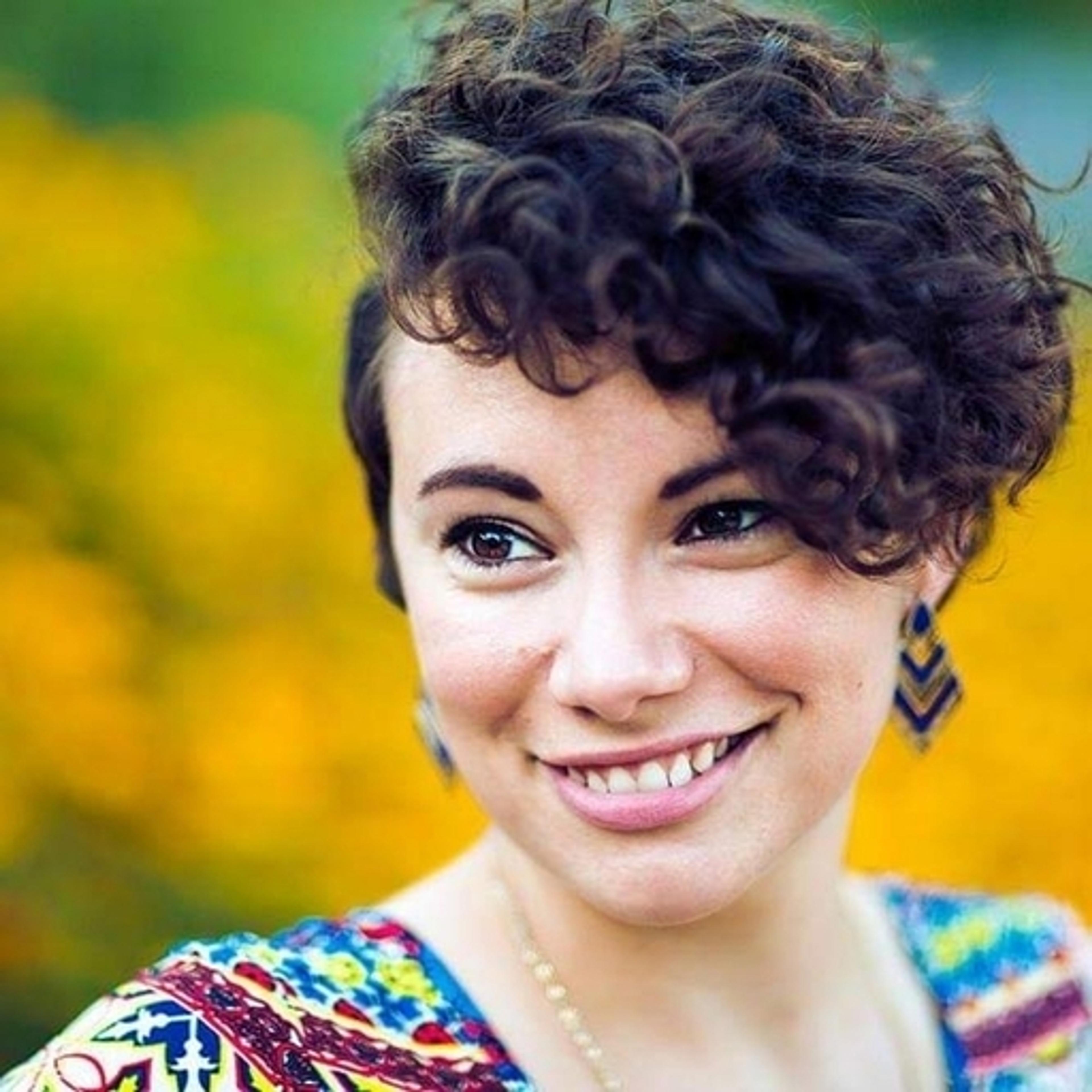 Mulher sorrindo com cabelo cacheado e curto, usando brincos e uma blusa colorida, em um fundo de flores amarelas.
