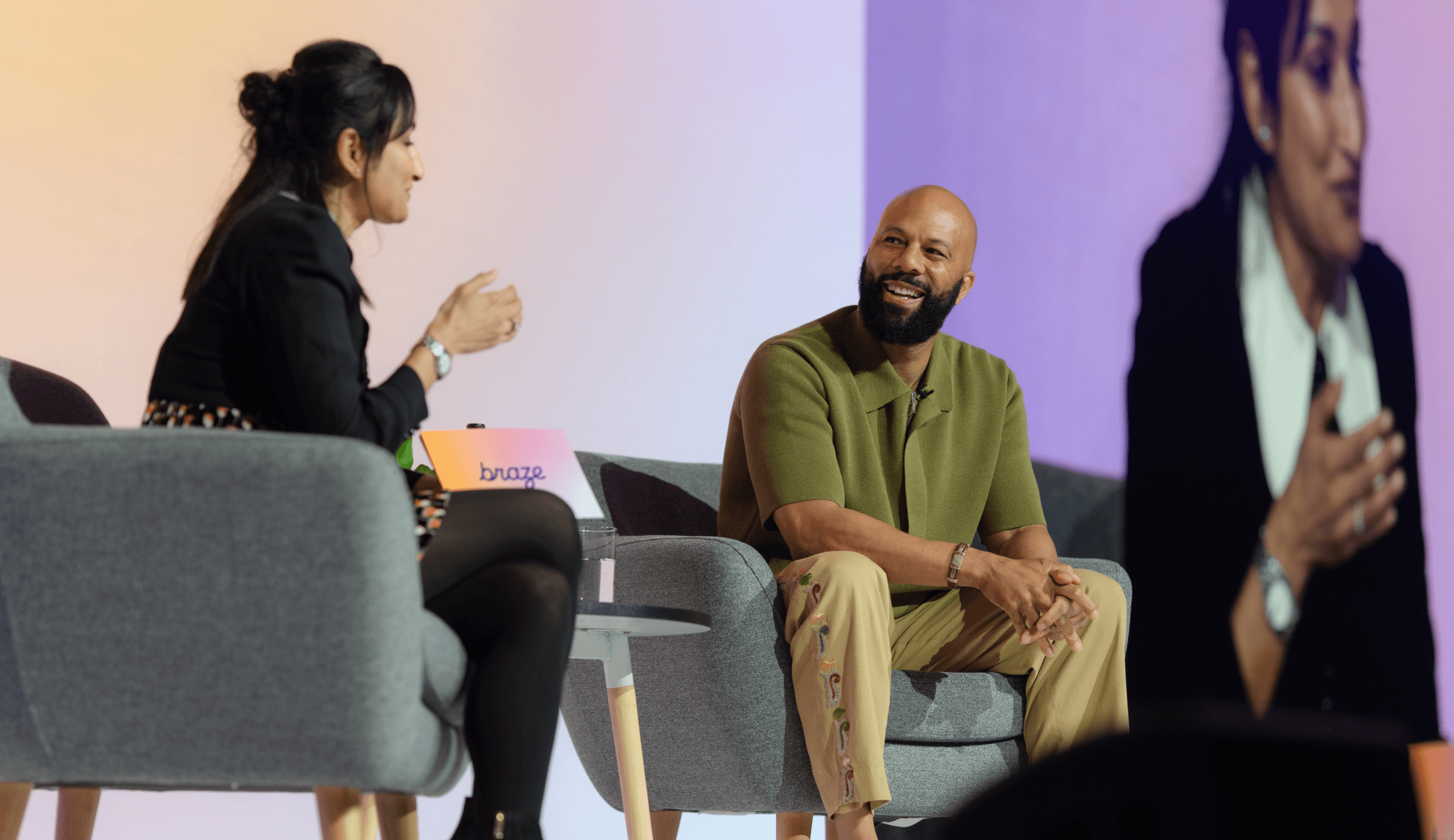 Two people sit in armchairs on a stage having a conversation, with one person smiling and a large screen behind them showing a close-up of the discussion.