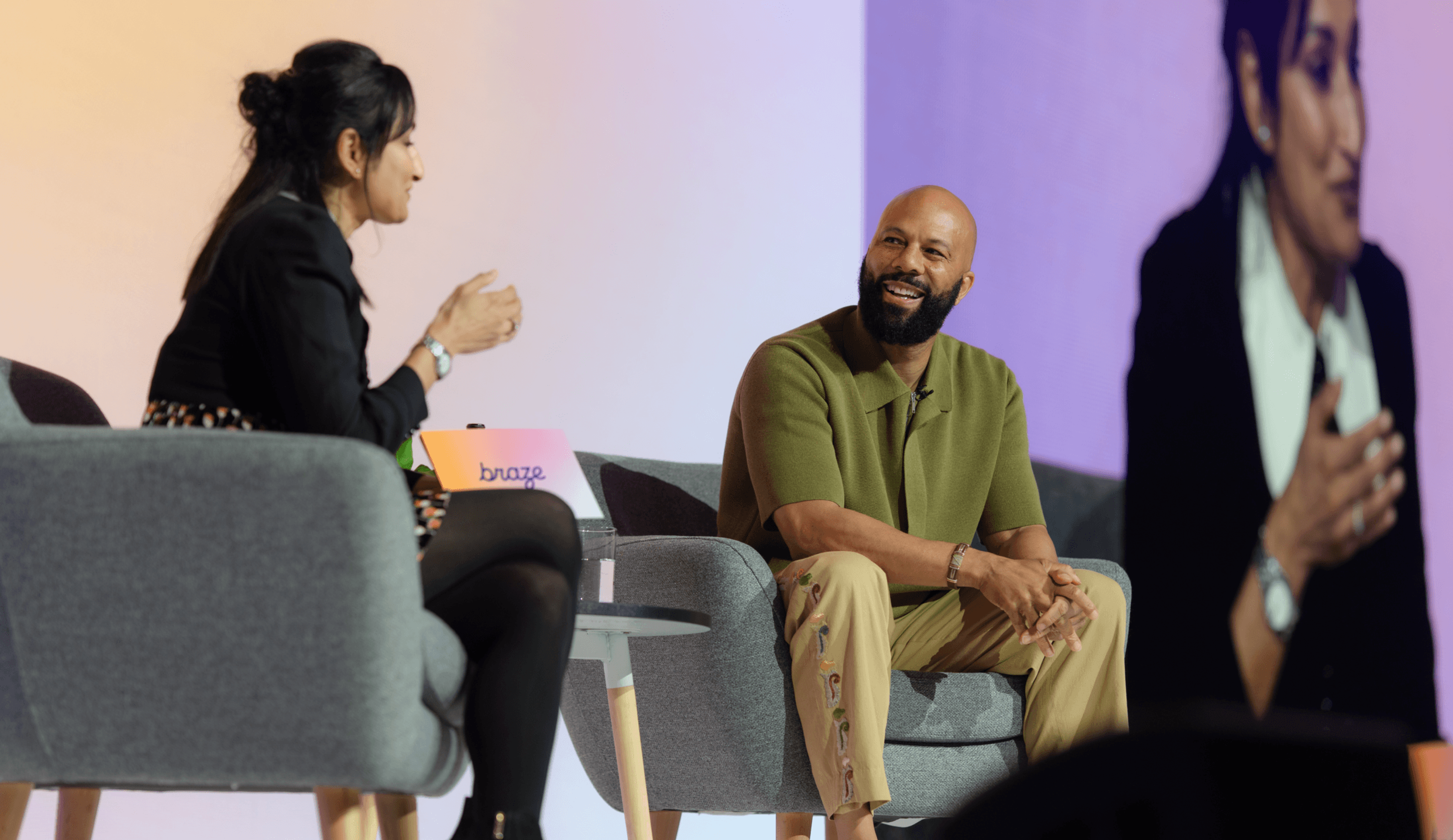 Two people sit in armchairs on a stage having a conversation, with one person smiling and a large screen behind them showing a close-up of the discussion.