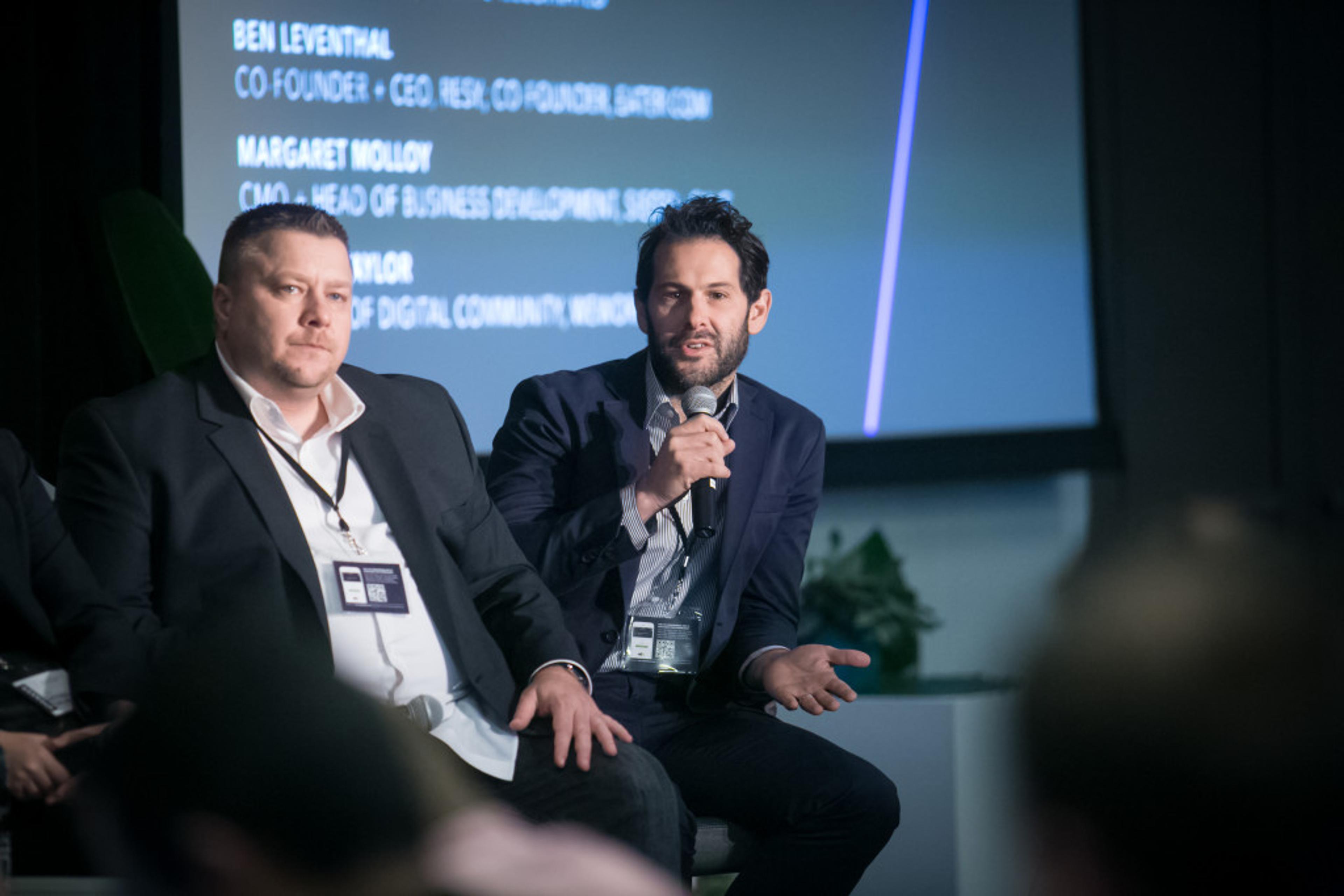 A panel discussion featuring two speakers, one actively speaking into a microphone while the other listens. A screen in the background displays their names and titles.