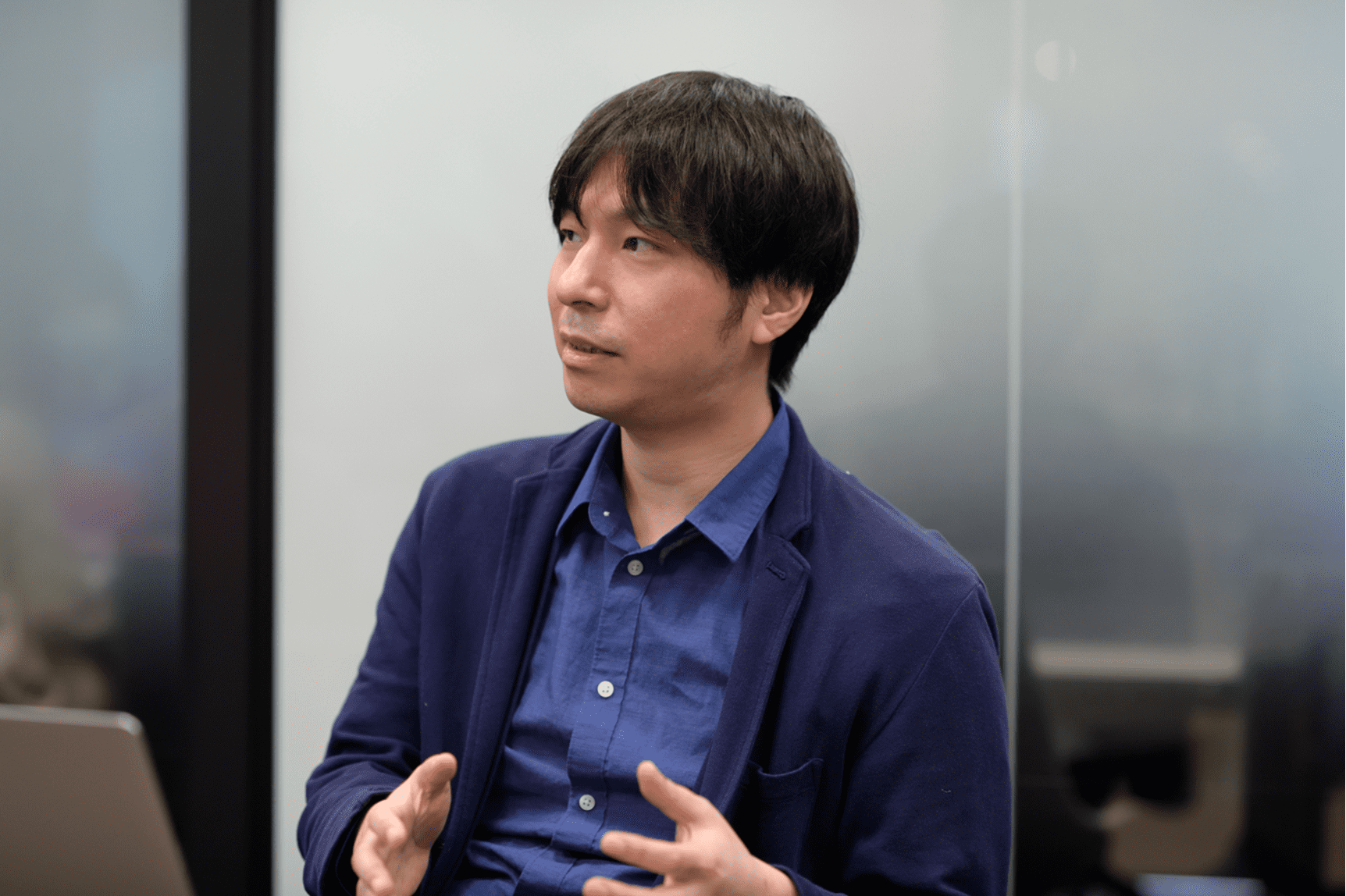 男性が会議室で話している様子。青いシャツとジャケットを着ています。