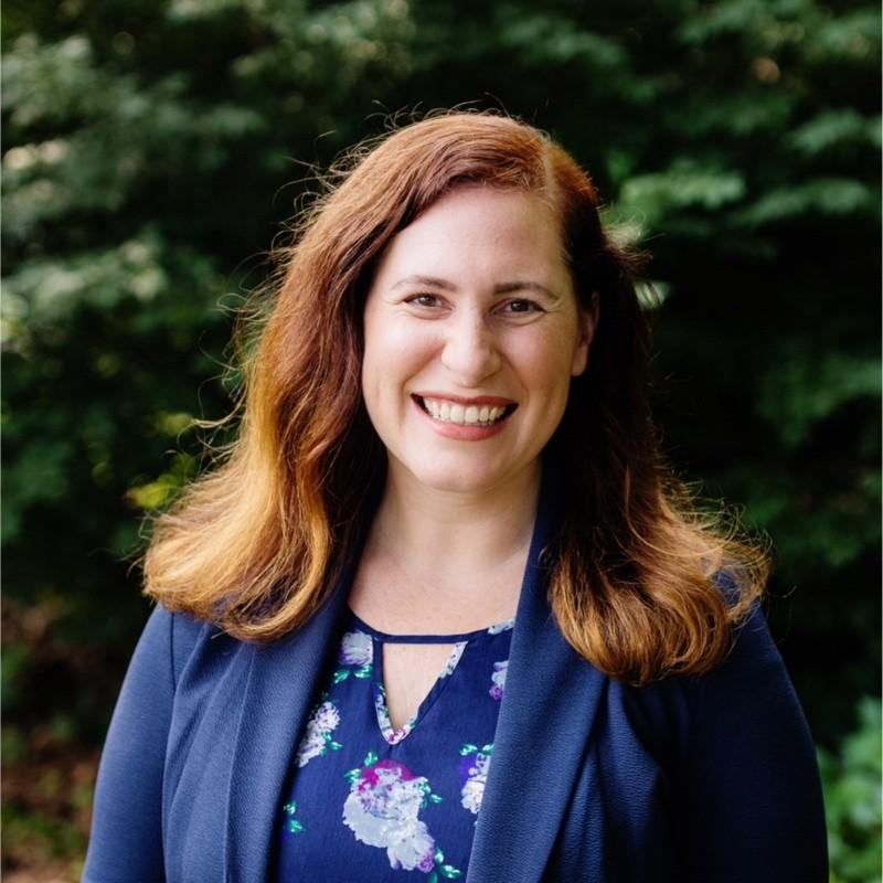 A woman with long, wavy brown hair smiles warmly at the camera, wearing a floral blouse and a navy blazer, with greenery in the background.