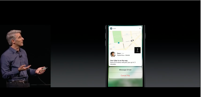 A man gestures while speaking next to a smartphone displaying a ride-sharing app interface, showing a driver's arrival time and options to message or cancel the trip.