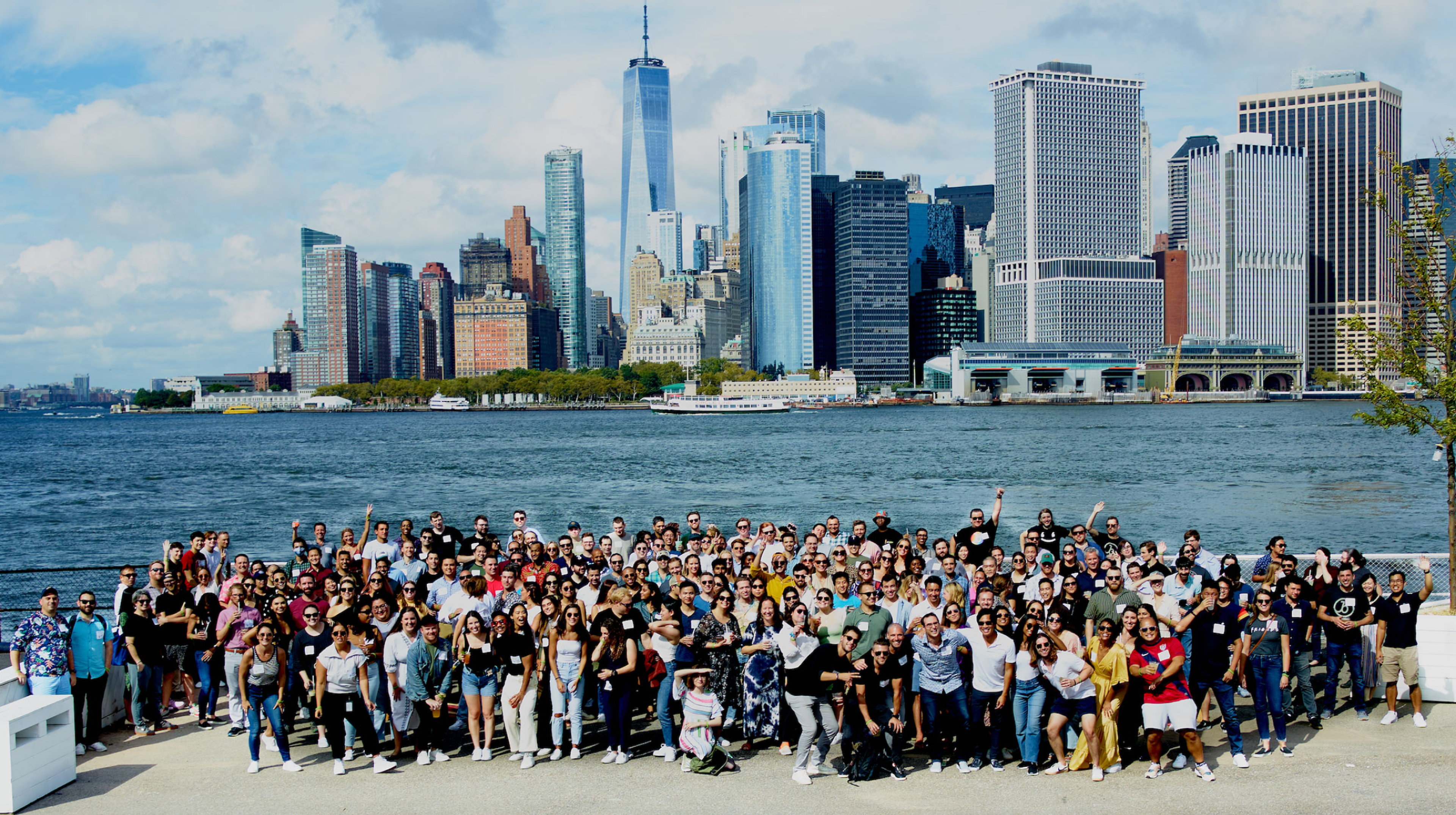 A large group of people poses for a photo in front of a city skyline with tall buildings and a body of water. The scene is bright and sunny, showcasing a vibrant urban environment.