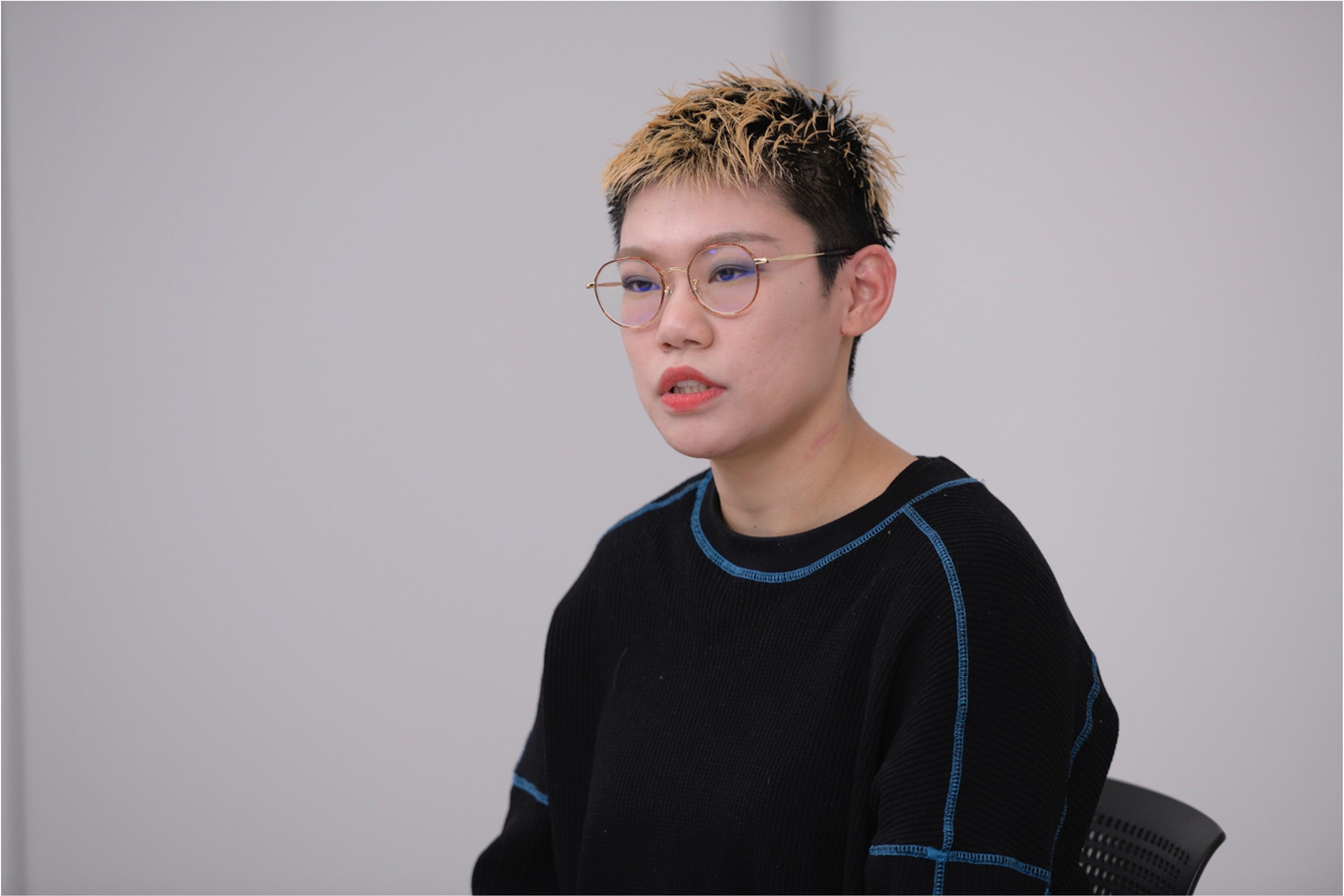 短髪で金髪の女性が黒いセーターを着て話しています。彼女はメガネをかけています。