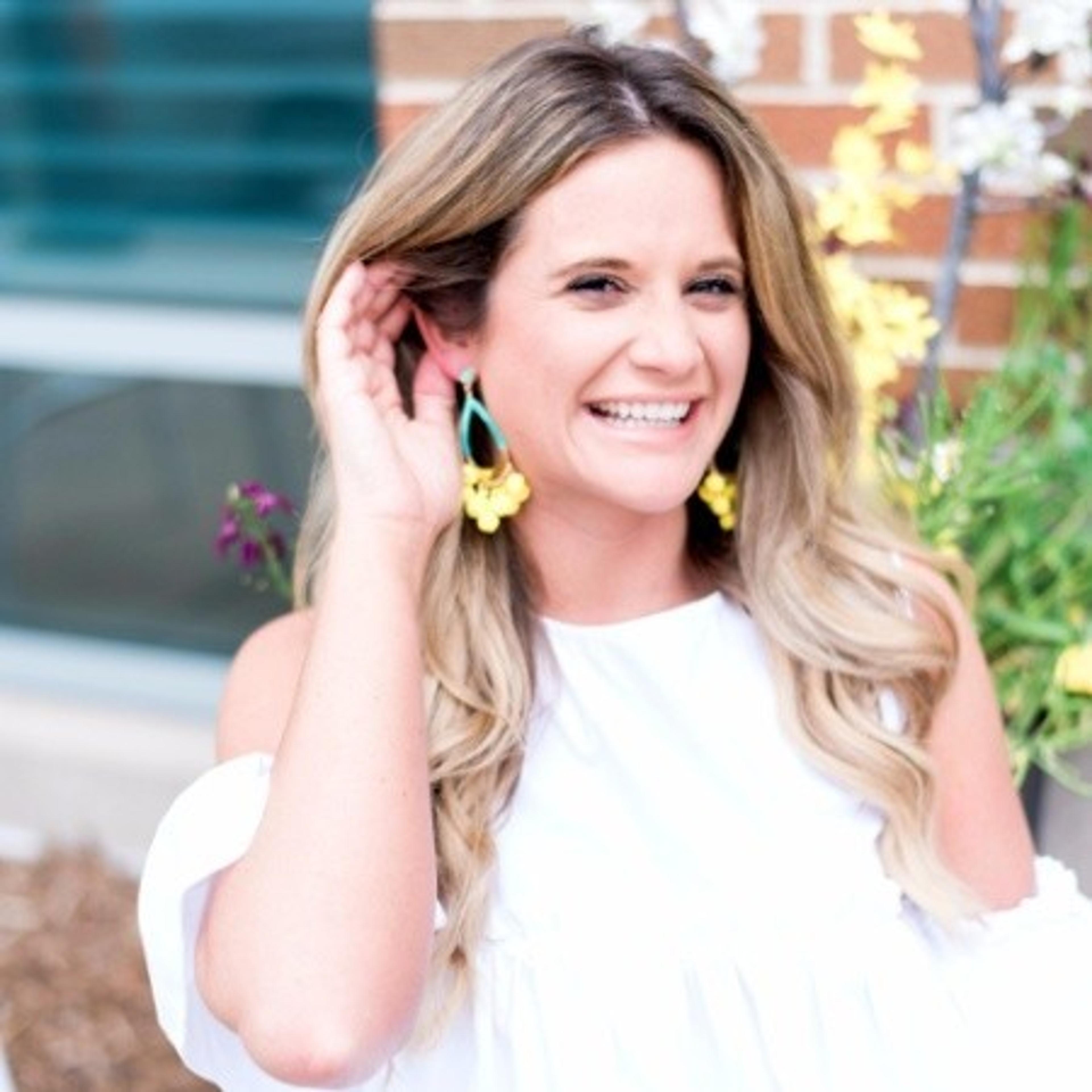 A woman with long, wavy hair smiles while touching her ear, wearing large yellow earrings. She stands in front of a floral backdrop.