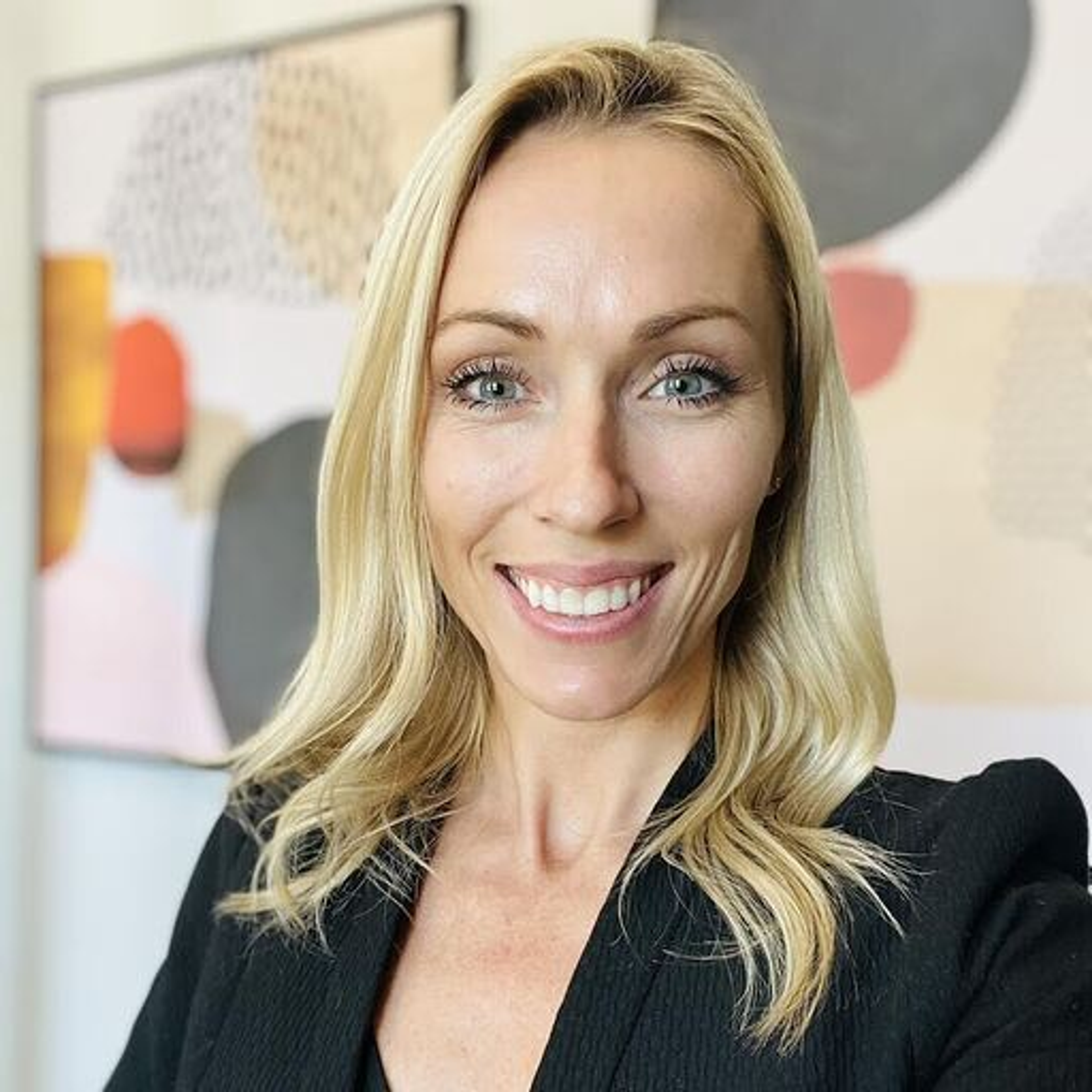 A woman with long blonde hair smiles at the camera, wearing a black blazer. Abstract art pieces in soft colors are visible in the background.