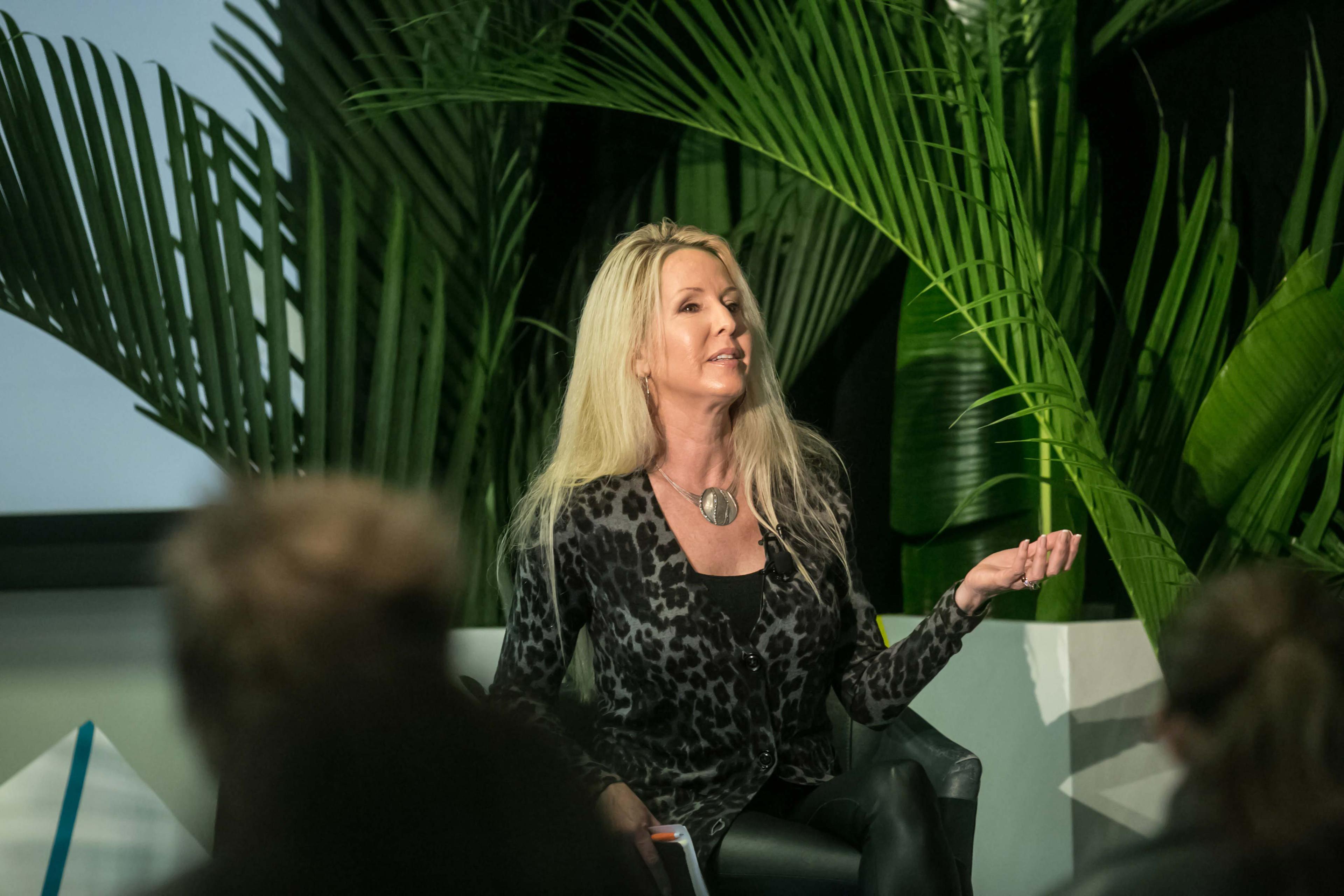 A woman with long blonde hair speaks while seated in front of large green plants. She wears a leopard print top and gestures with her hand.