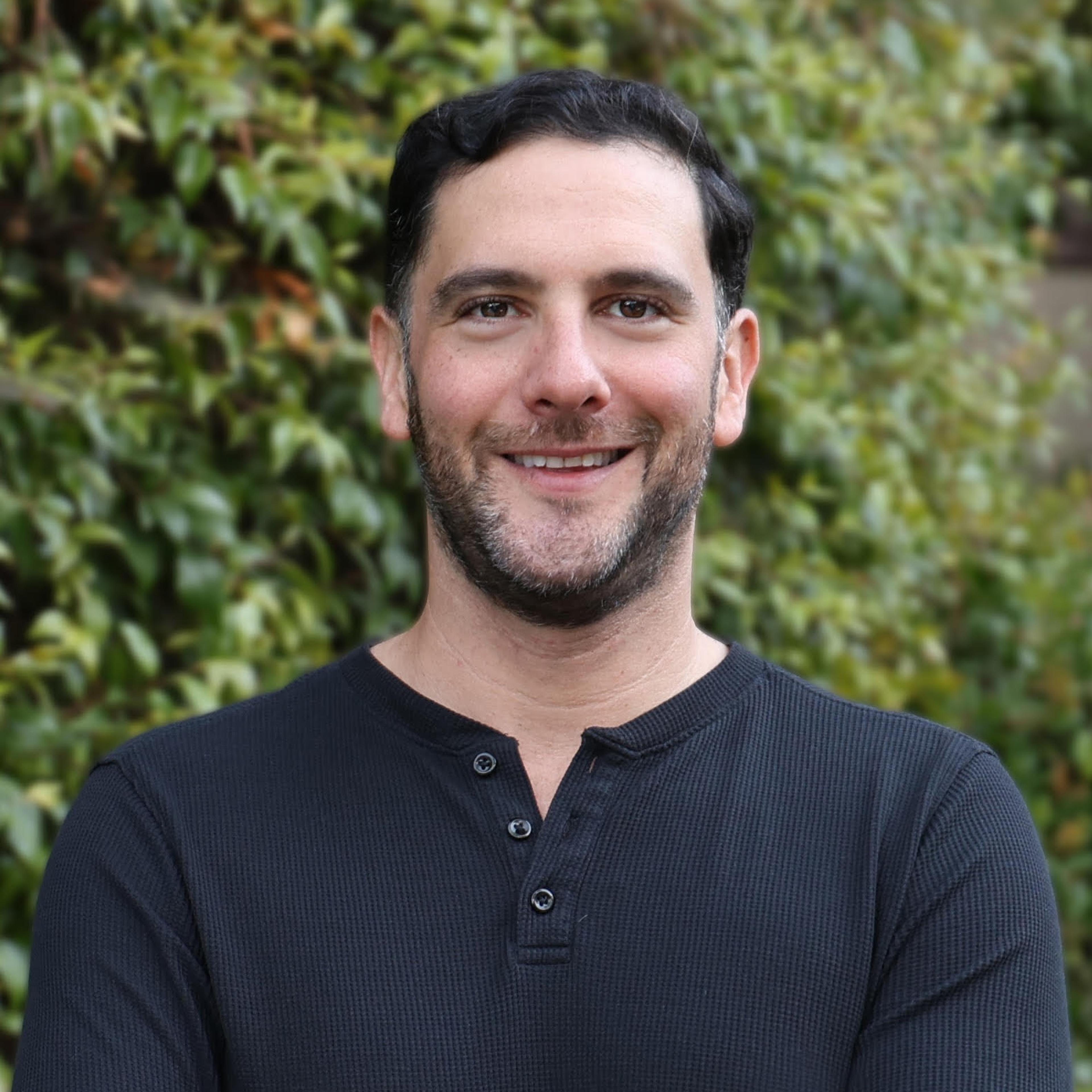 A smiling man with dark hair and a beard stands with his arms crossed in front of a green leafy background. He is wearing a black long-sleeve shirt.