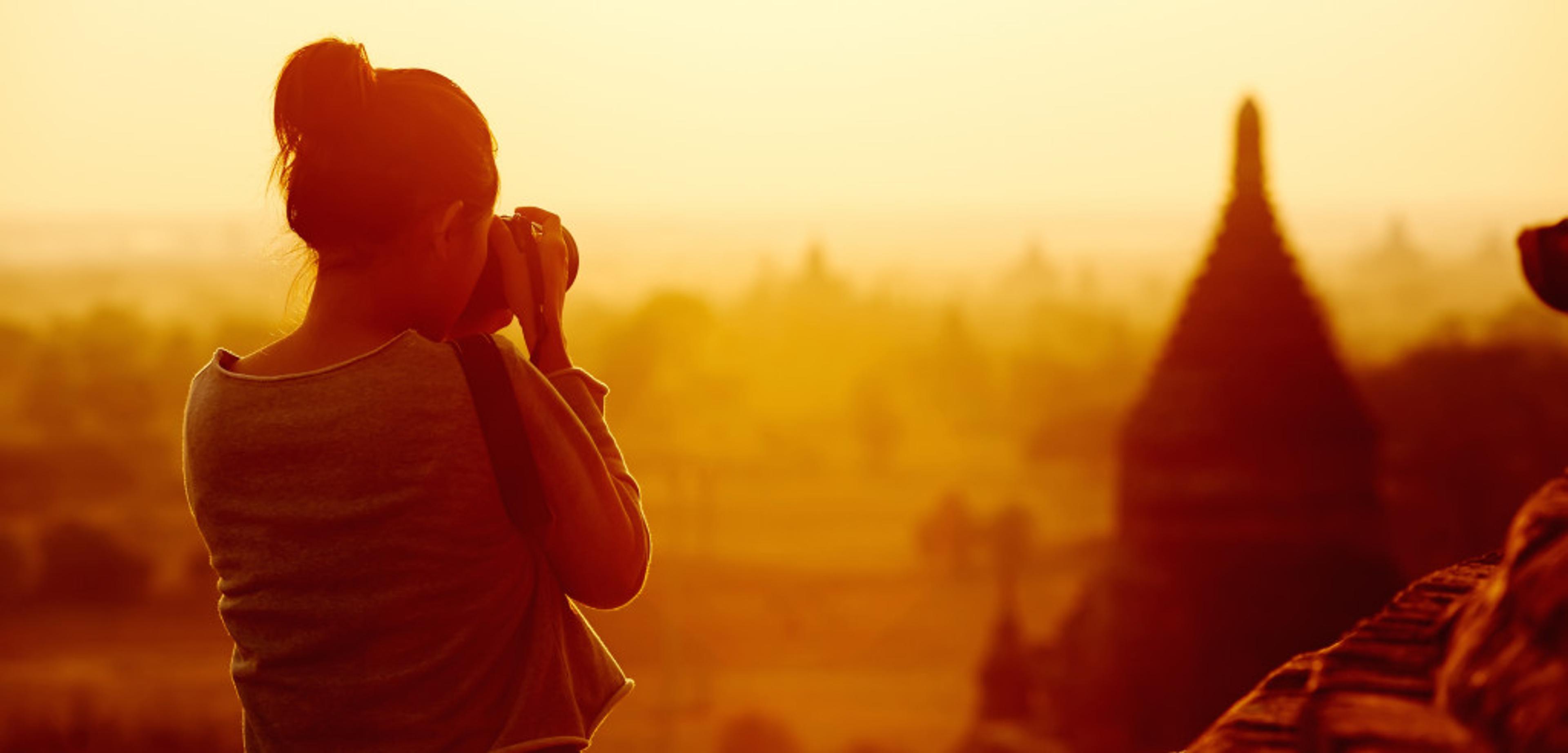 A person stands silhouetted against a golden sunset, holding a camera and looking out over a landscape with pagoda-like structures. The scene captures a serene moment in a picturesque setting.