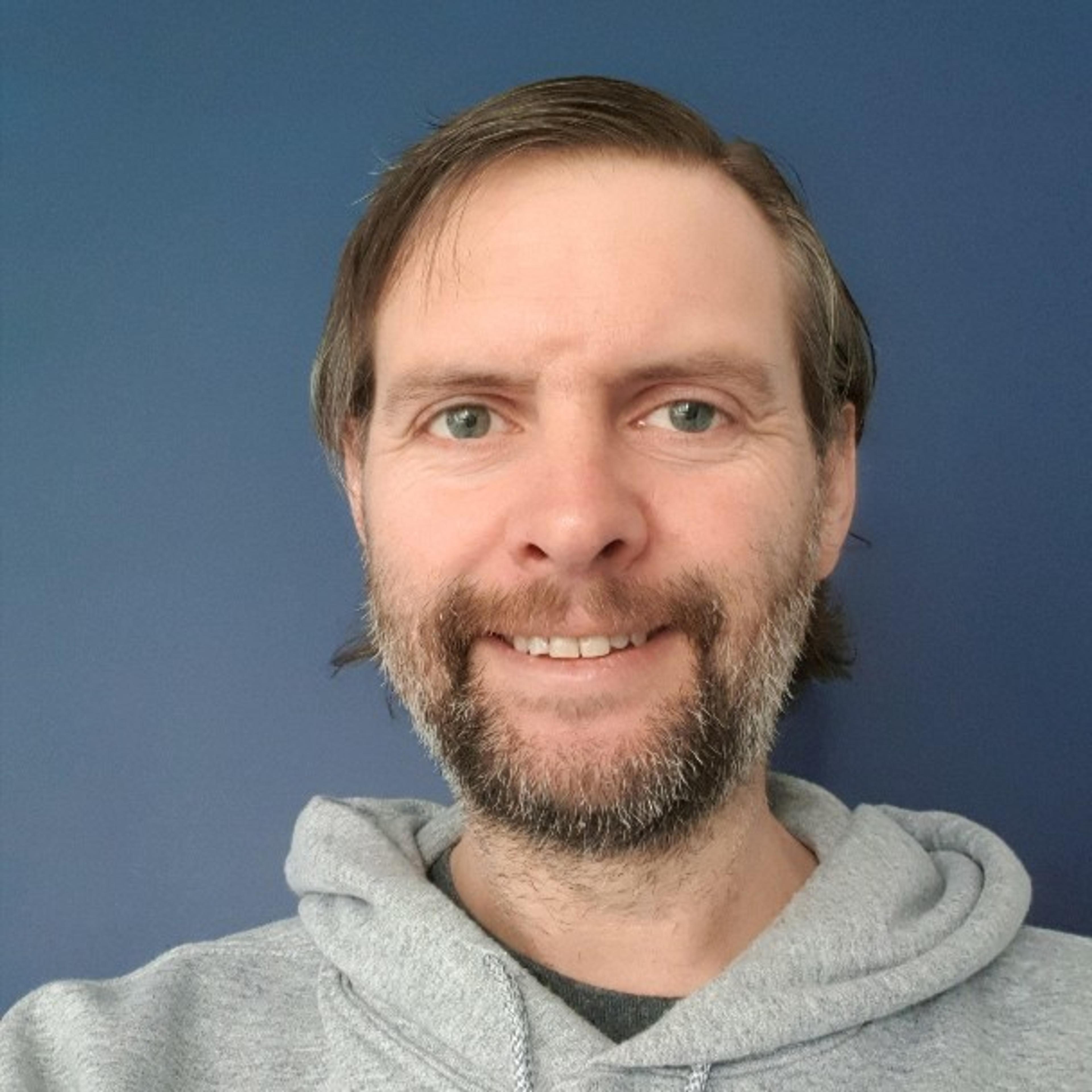 Smiling bearded person in a gray hoodie standing in front of a plain dark blue wall.
