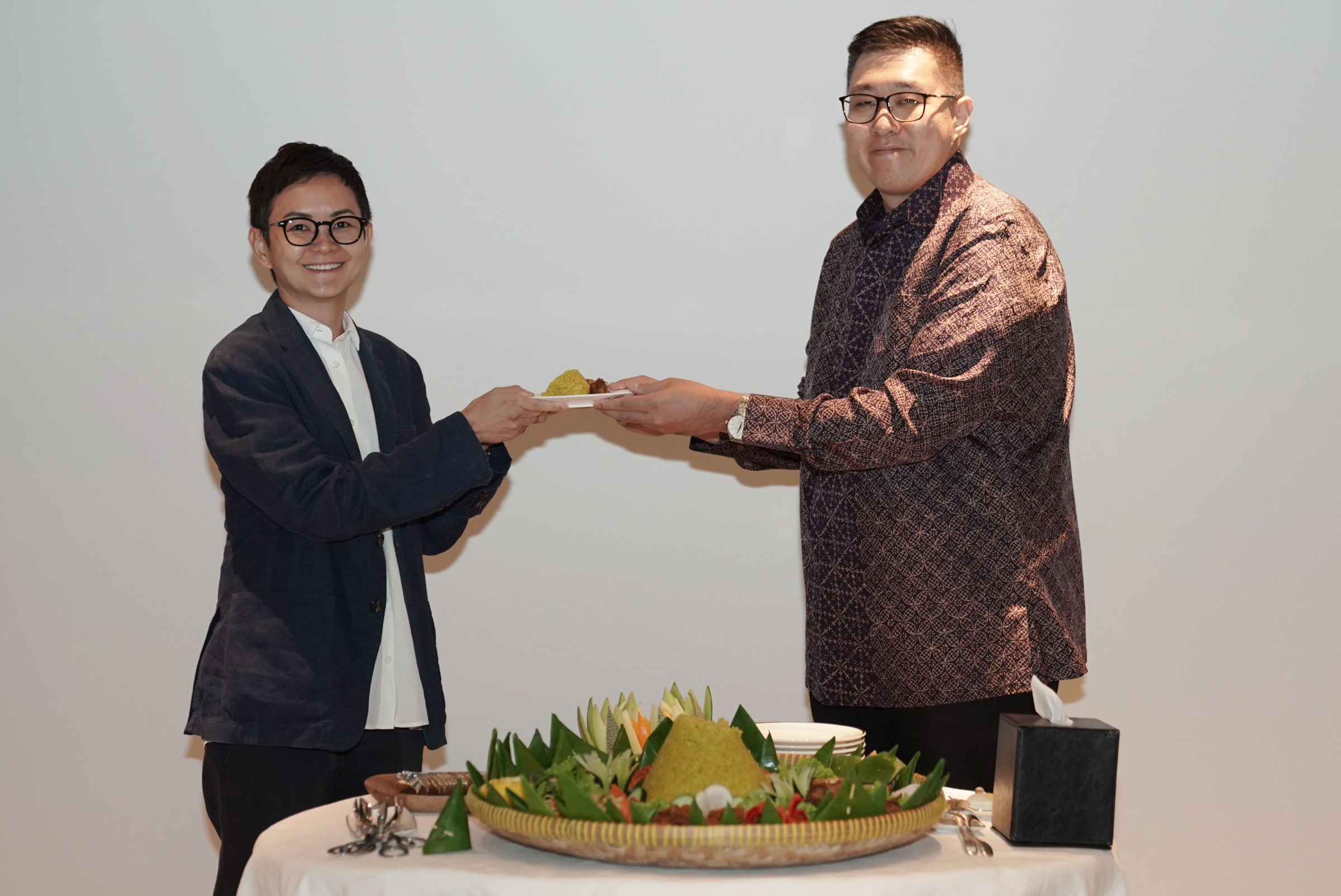 Two people are standing side by side, each holding a plate with a serving of food. A decorative platter with rice and vegetables is displayed on a table in front of them.