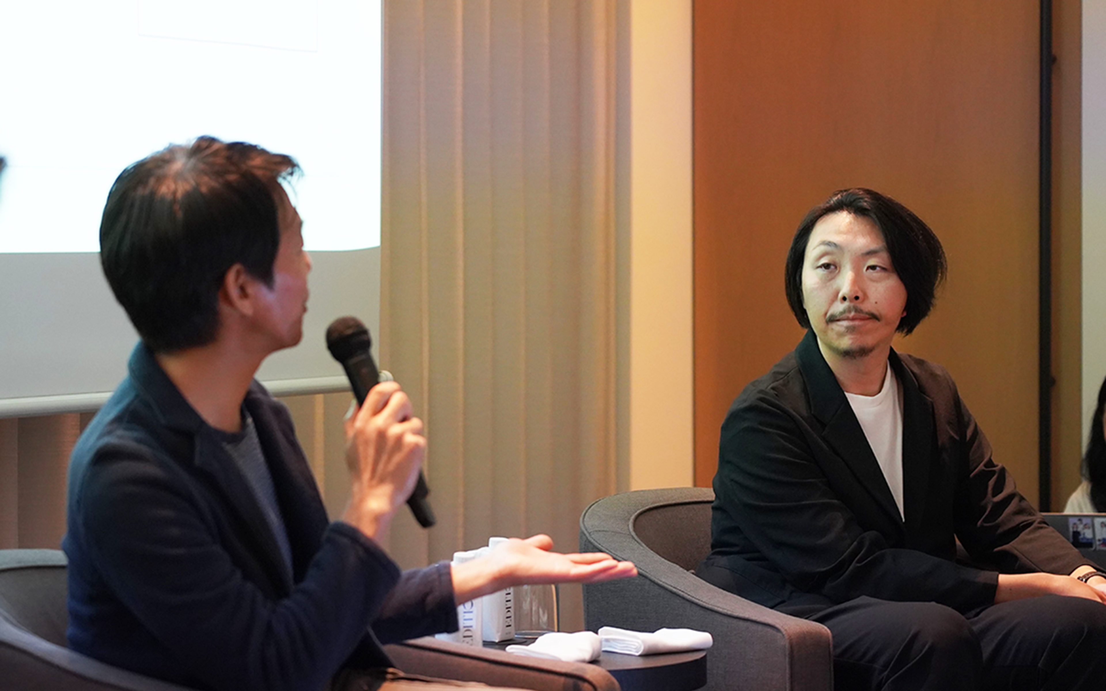 二人の男性が座って対話をしています。一人はマイクを持ち、話している様子です。