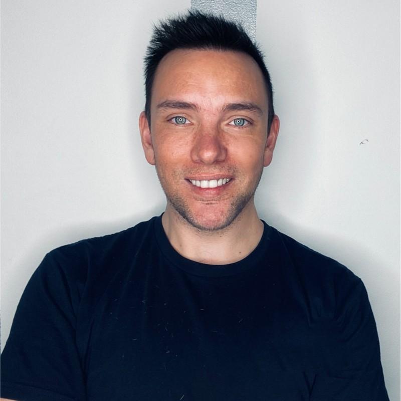 Headshot of a smiling man with short dark hair and blue eyes, wearing a black shirt against a white background.