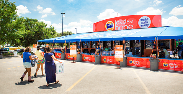 A colorful Tide Loads of Hope mobile laundry service set up outdoors, with people walking towards it. The tent features washing machines and a bright sign promoting the service.