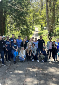 A group of people poses together on a paved path in a sunlit wooded park.