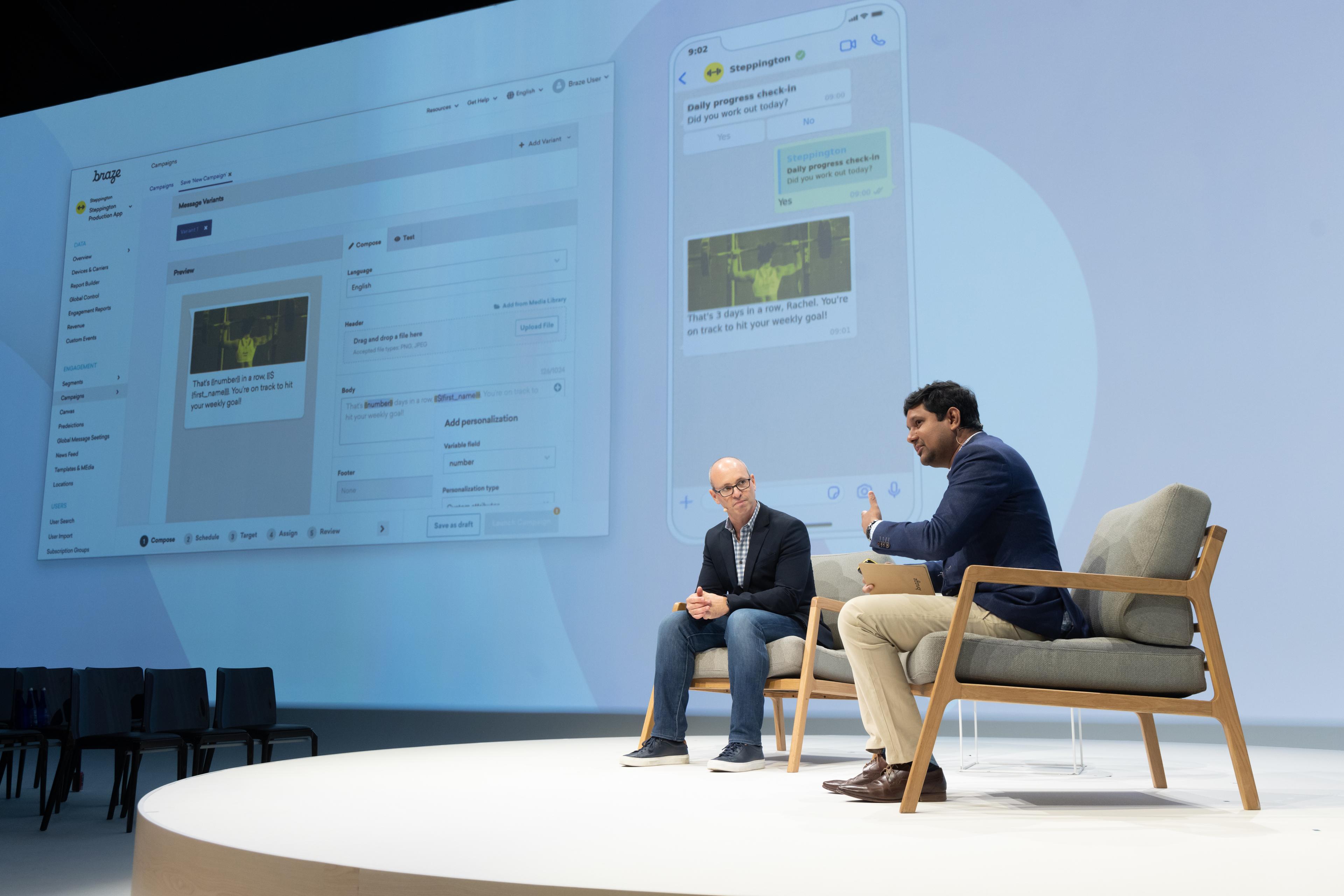 Two men sit on a stage discussing a digital interface displayed on a large screen behind them. One man is speaking while the other listens attentively.