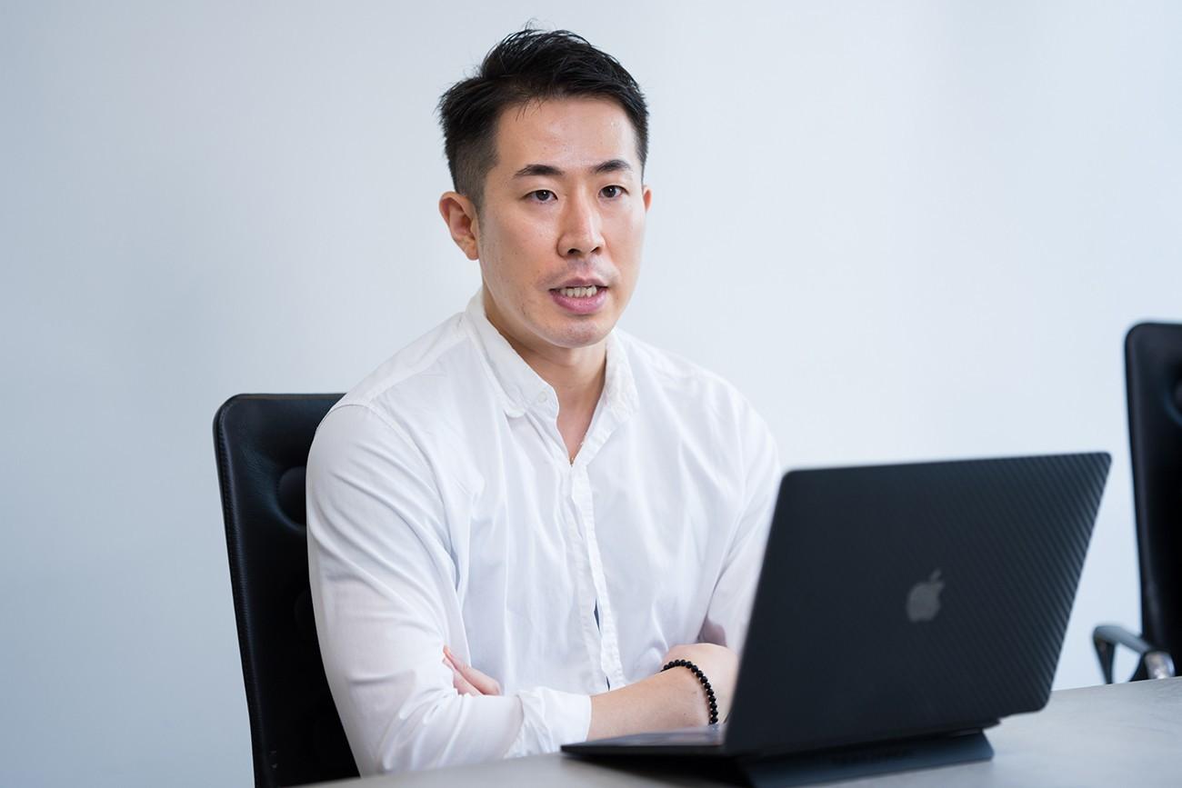 男性がノートパソコンの前で話している様子。白いシャツを着ており、真剣な表情をしています。