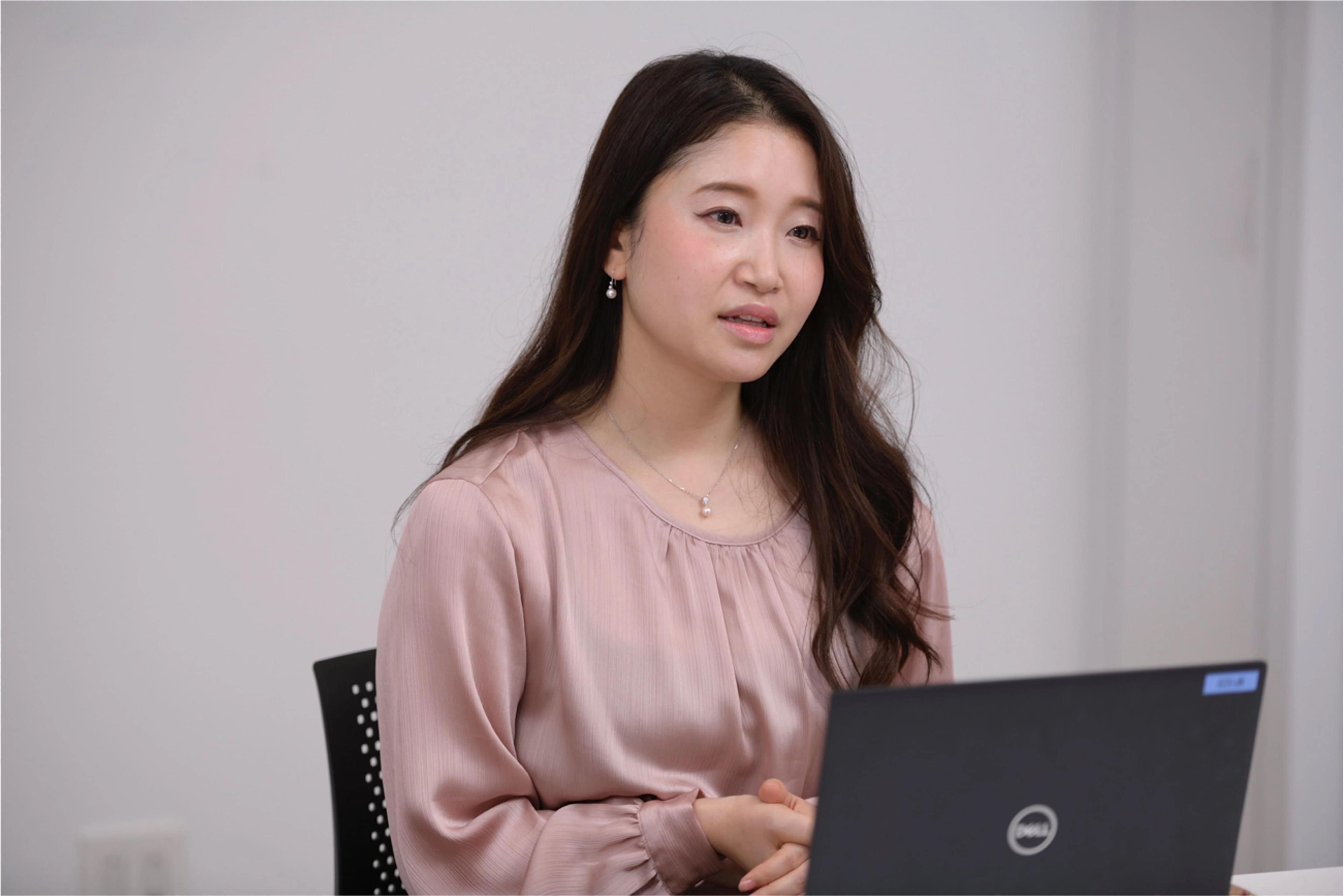 女性がノートパソコンの前で話している様子。彼女は薄いピンクのブラウスを着ています。