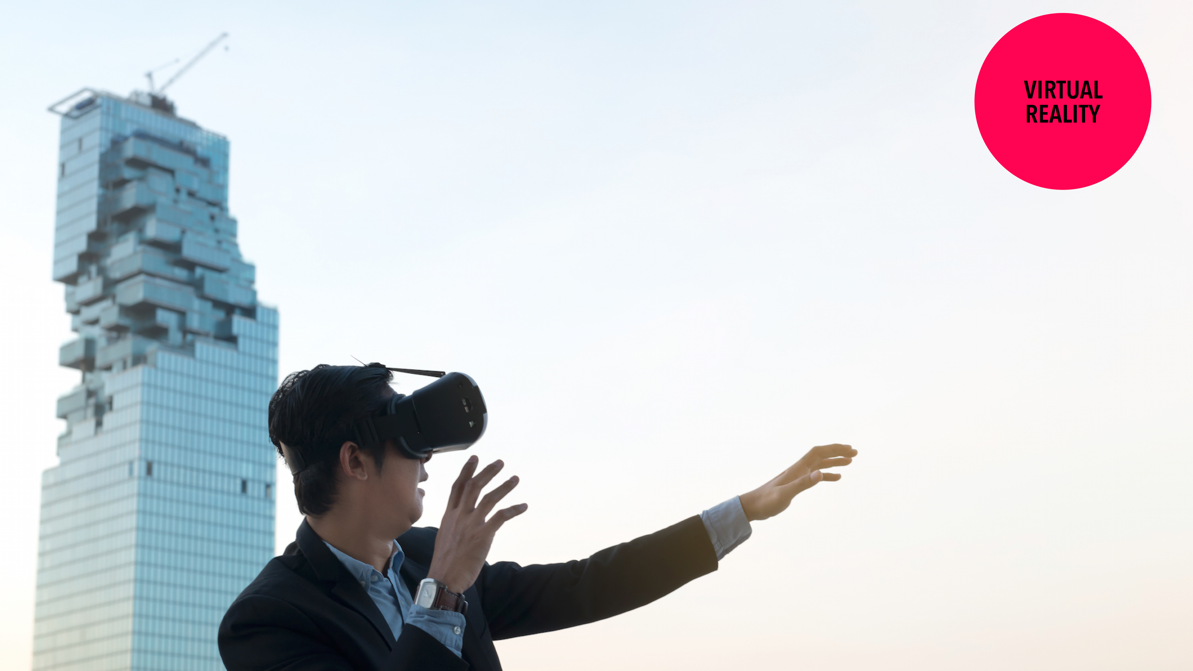 A man in a suit is using a virtual reality headset outdoors, gesturing with one hand. A modern building is visible in the background.