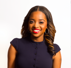 A smiling woman with long, wavy hair wearing a dark dress and hoop earrings. She has bright red lipstick and is posed against a white background.