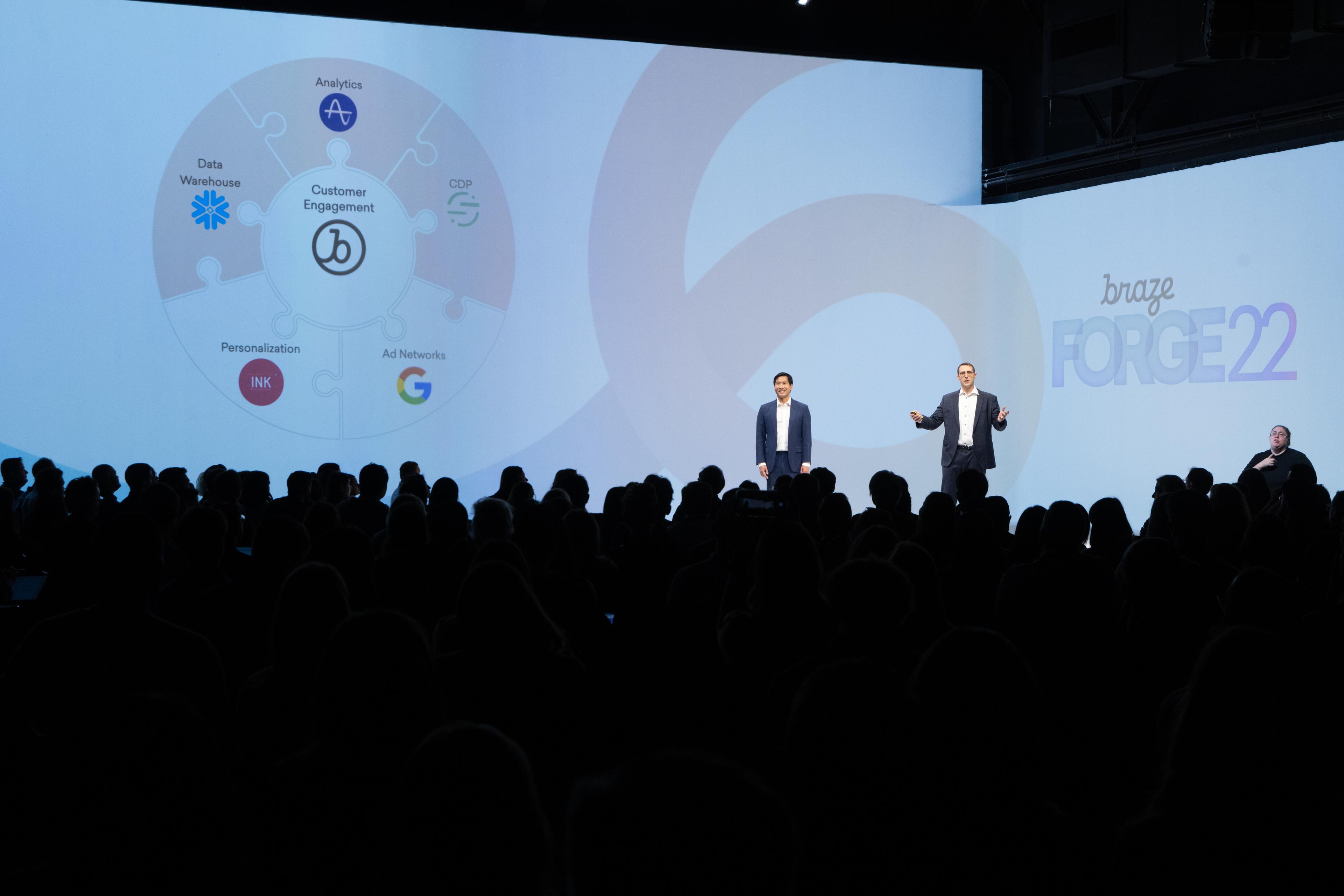 Two speakers on stage at a conference, presenting to an audience. A visual display behind them features a circular diagram related to analytics and customer engagement.