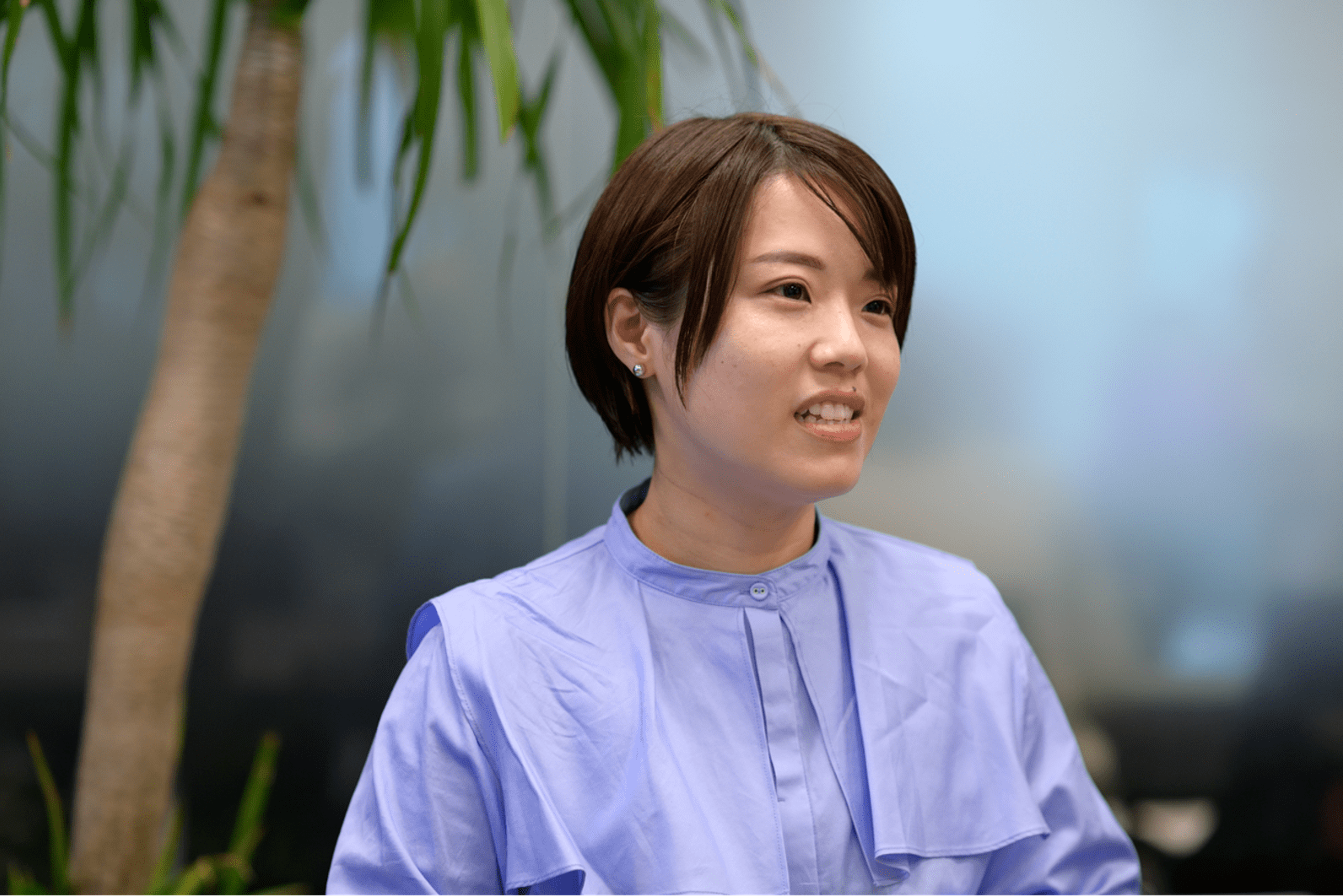 短い髪の女性が青いシャツを着て、植物の近くで微笑んでいます。背景はぼやけています。