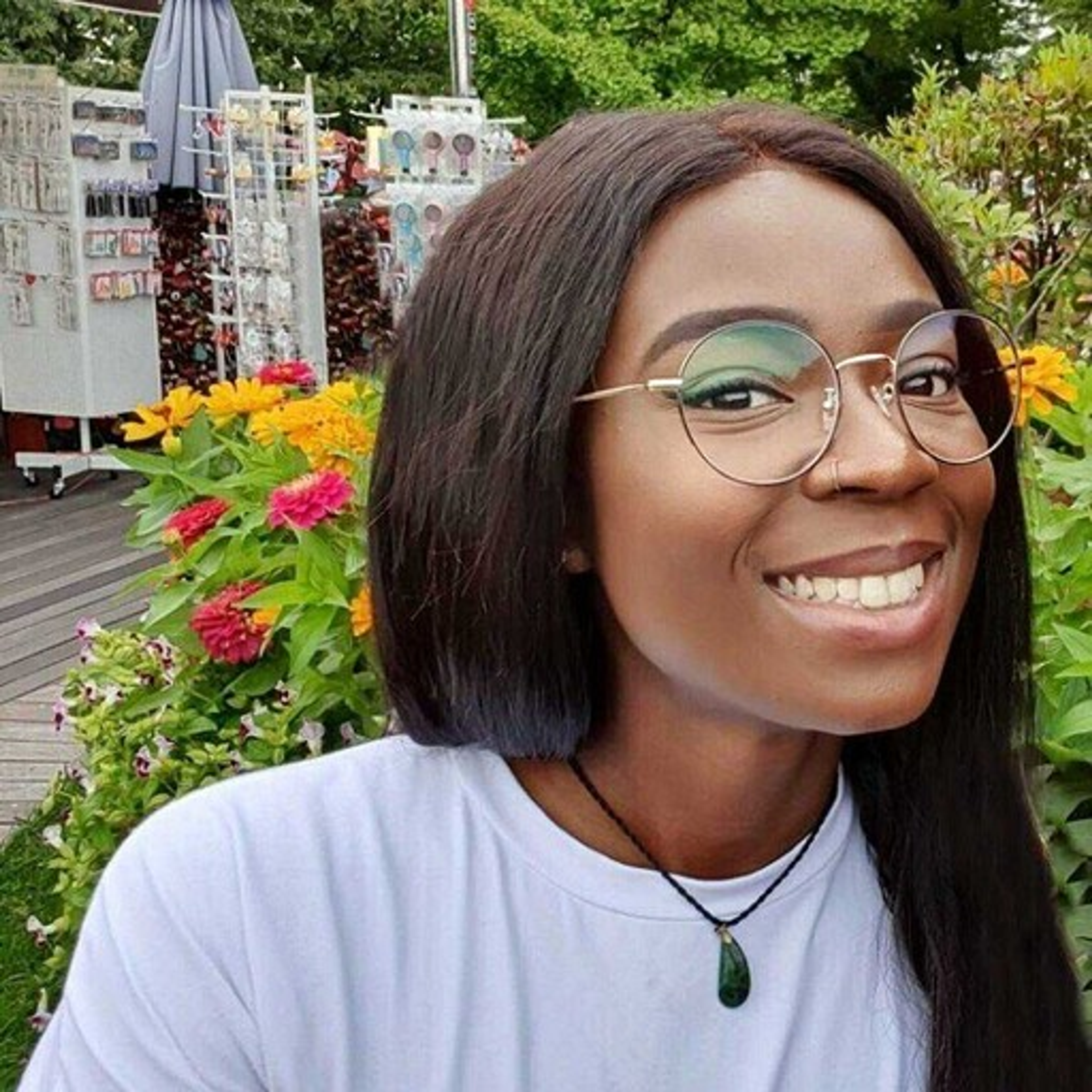 A smiling person with long dark hair and glasses poses in front of colorful flowers and a market stall. The background features various items for sale.